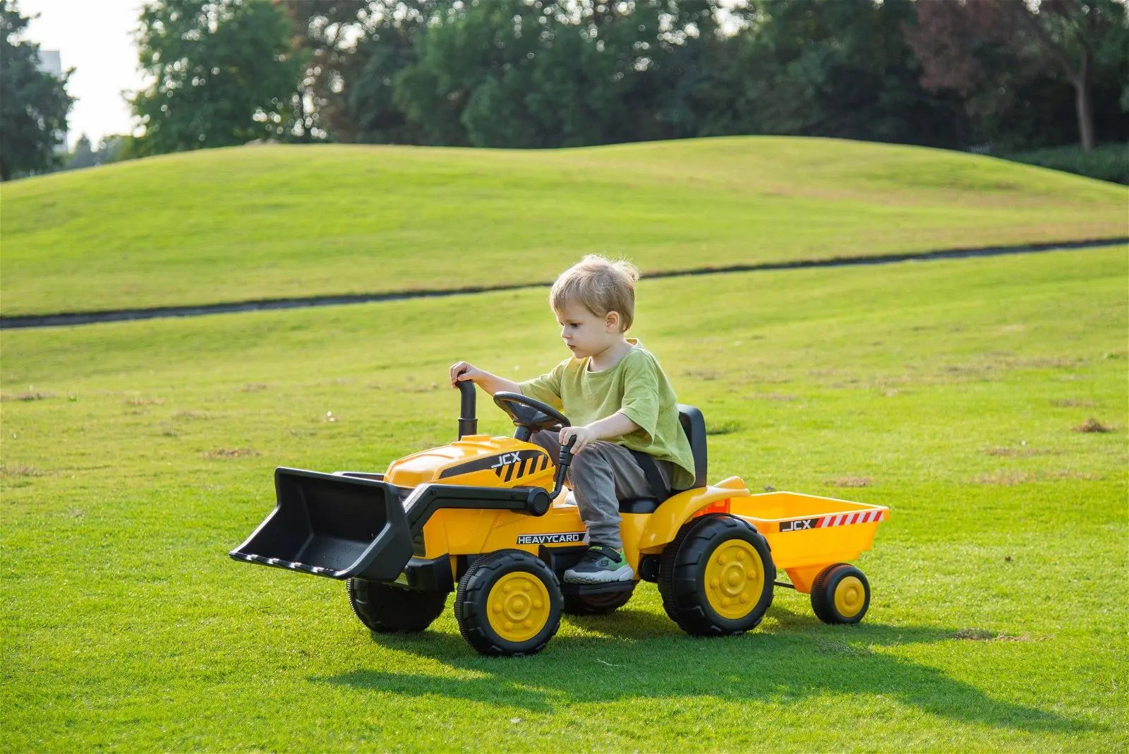 Child riding yellow 12V ride-on excavator toy with trailer on grassy outdoor field