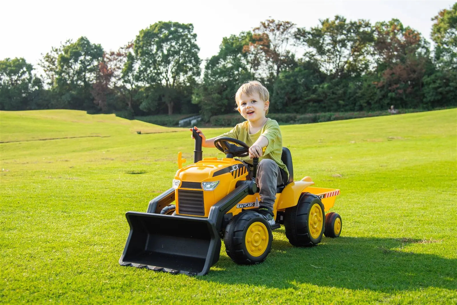 Child riding yellow 12V excavator toy vehicle outdoors on green grass field
