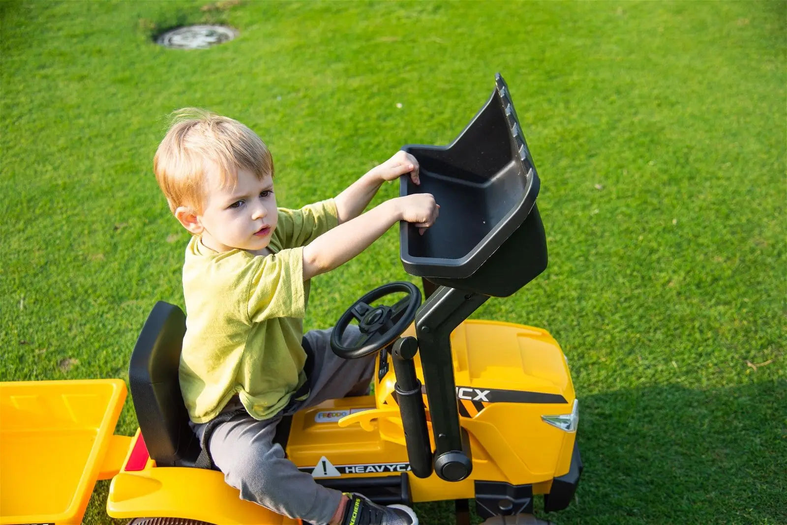 Child riding a yellow 12V toy excavator on green grass outdoors
