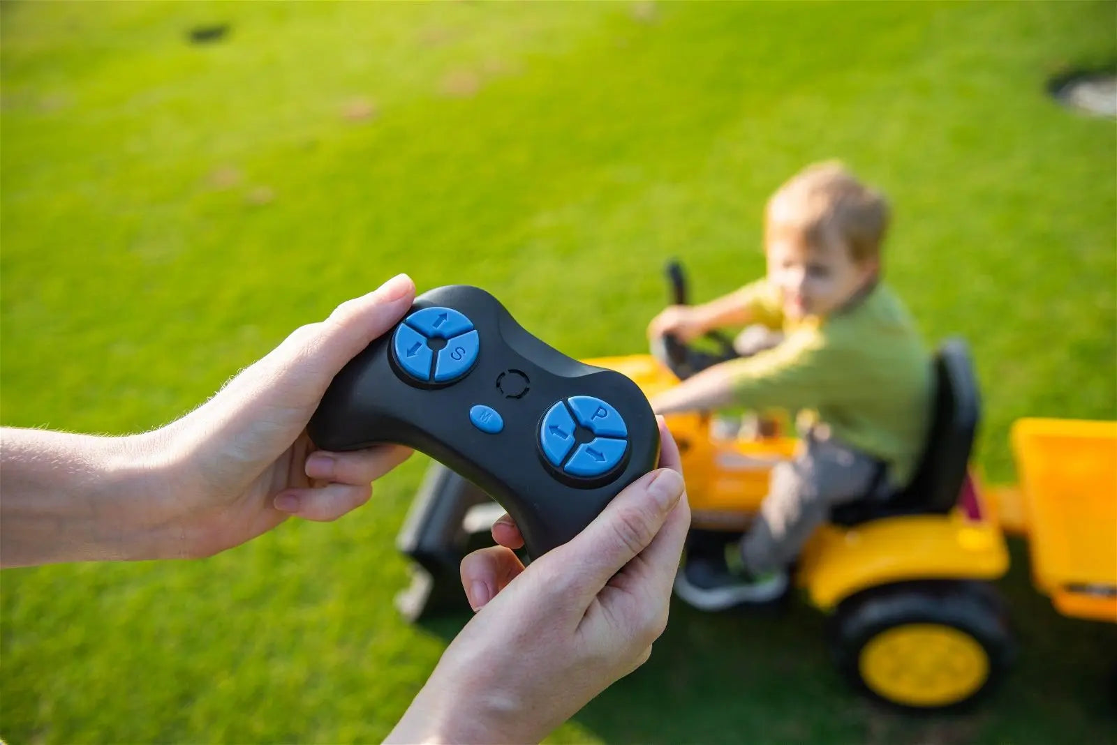 Hands holding remote control for kids ride-on excavator with child driving on green grass
