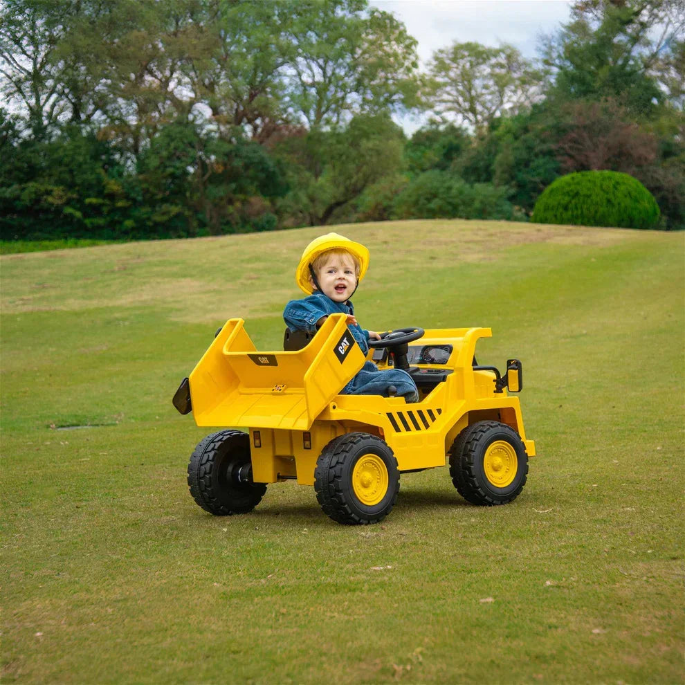 Toddler wearing a yellow helmet driving yellow 12V CAT electric dump truck ride-on toy in a grassy outdoor area
