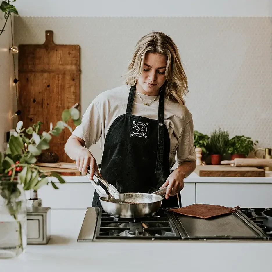 Woman in black apron cooking with tongs in a modern kitchen with plants and cutting boards.