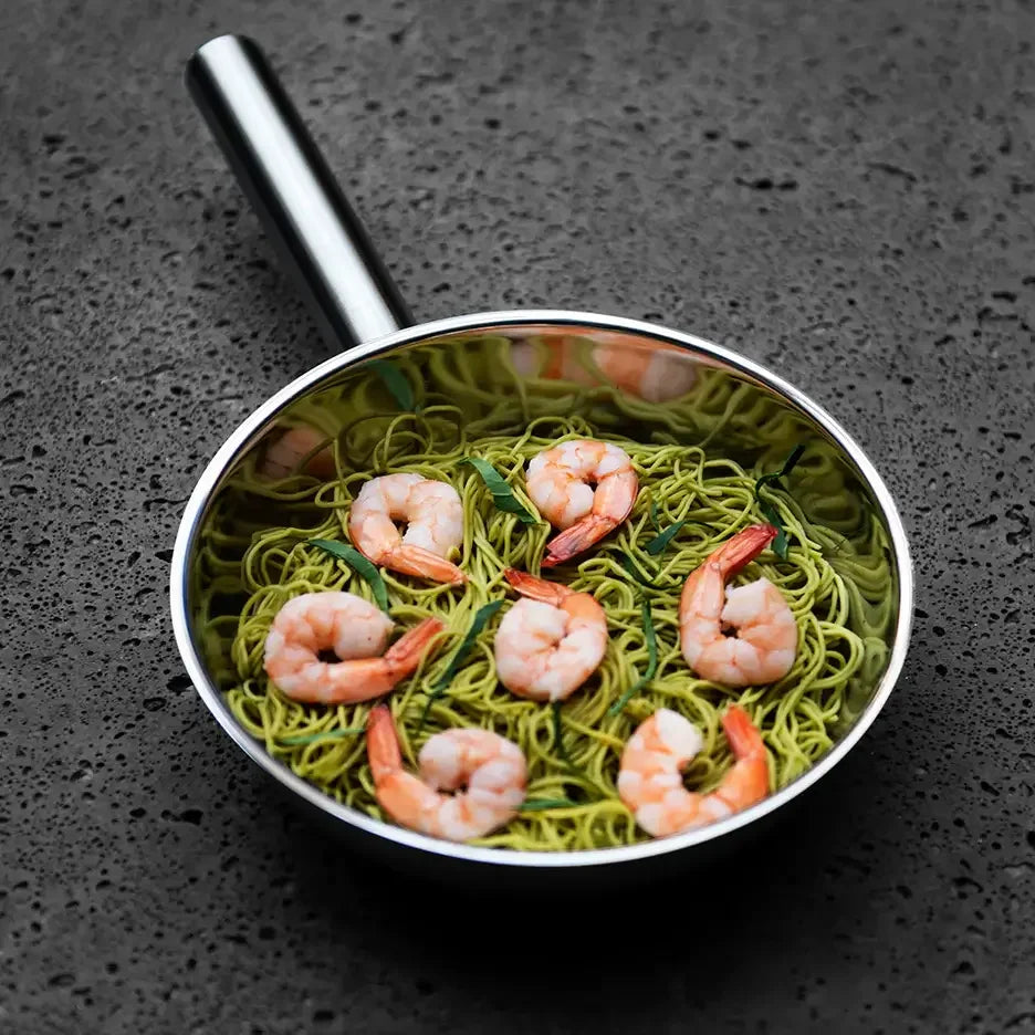 Stainless steel pan with shrimp and green noodles on a dark textured surface