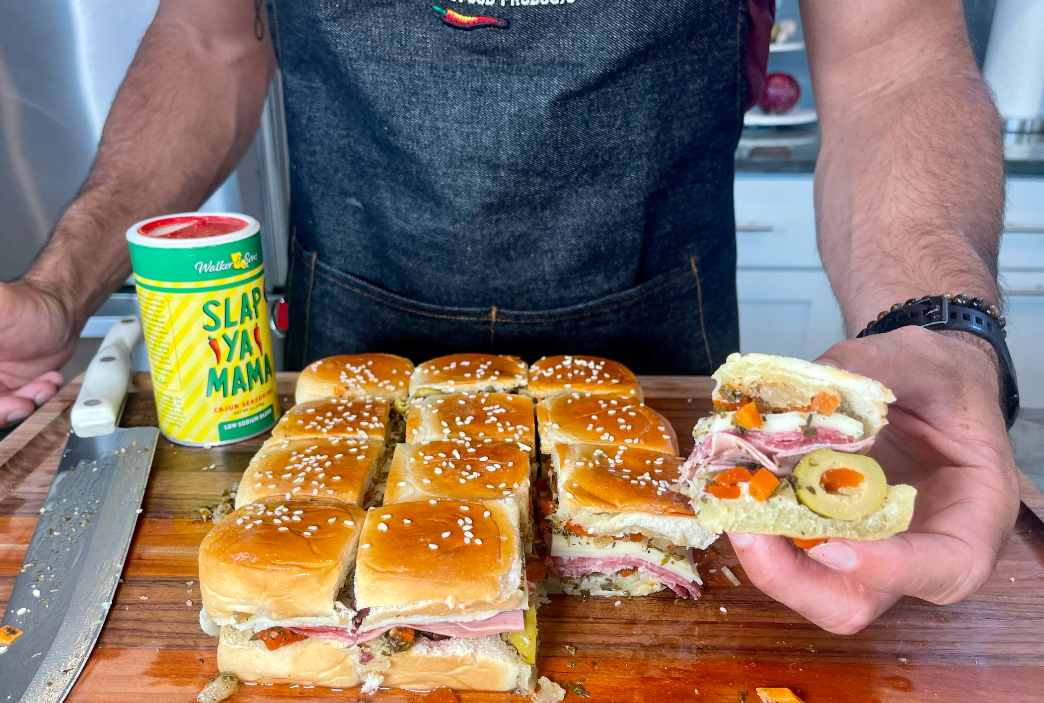 Person holding slider sandwich with deli meat, cheese, olives on cutting board, Cajun seasoning nearby