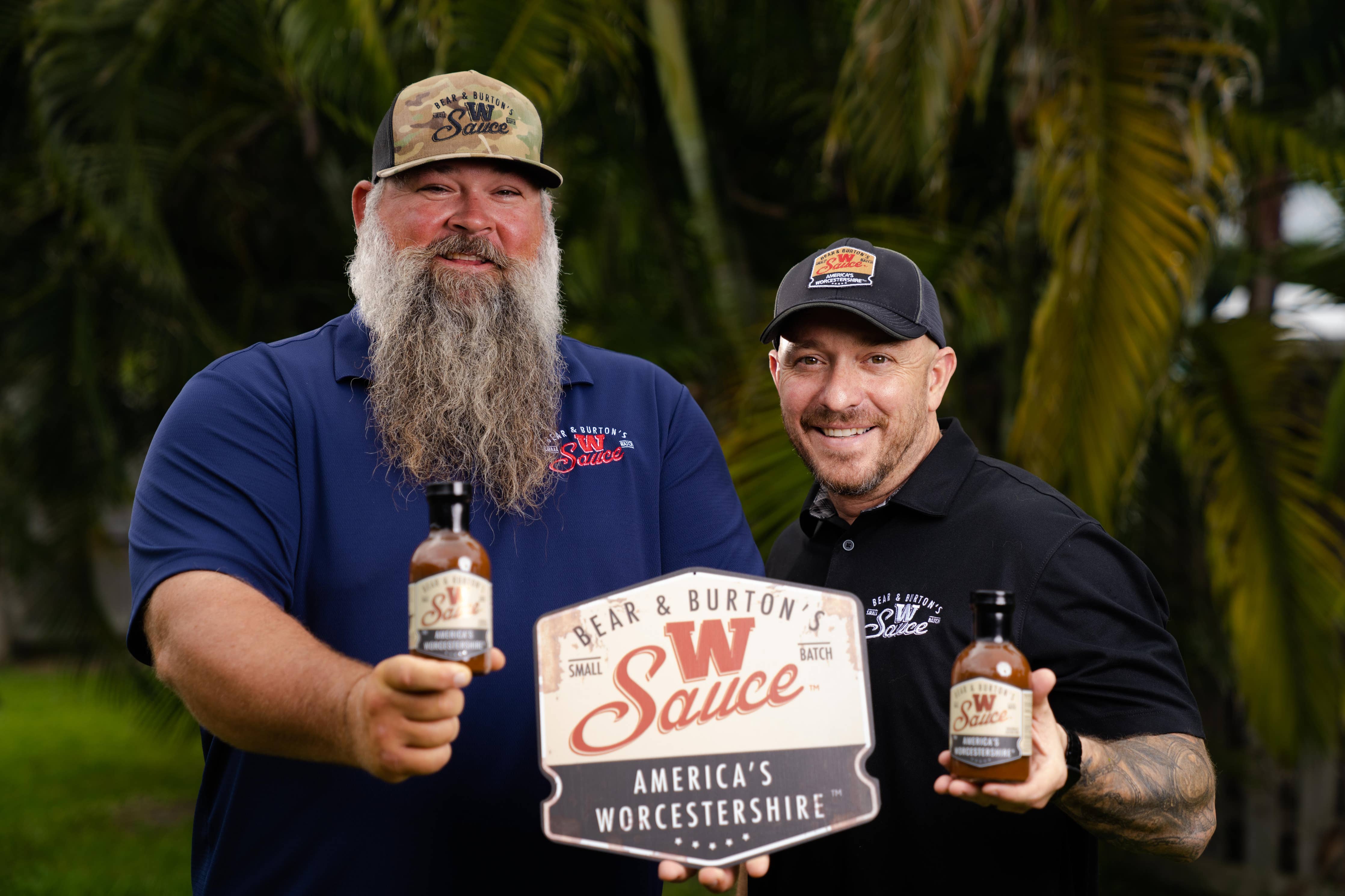 Two men outdoors holding Bear & Burton's W Sauce Worcestershire bottles and sign, palm trees behind