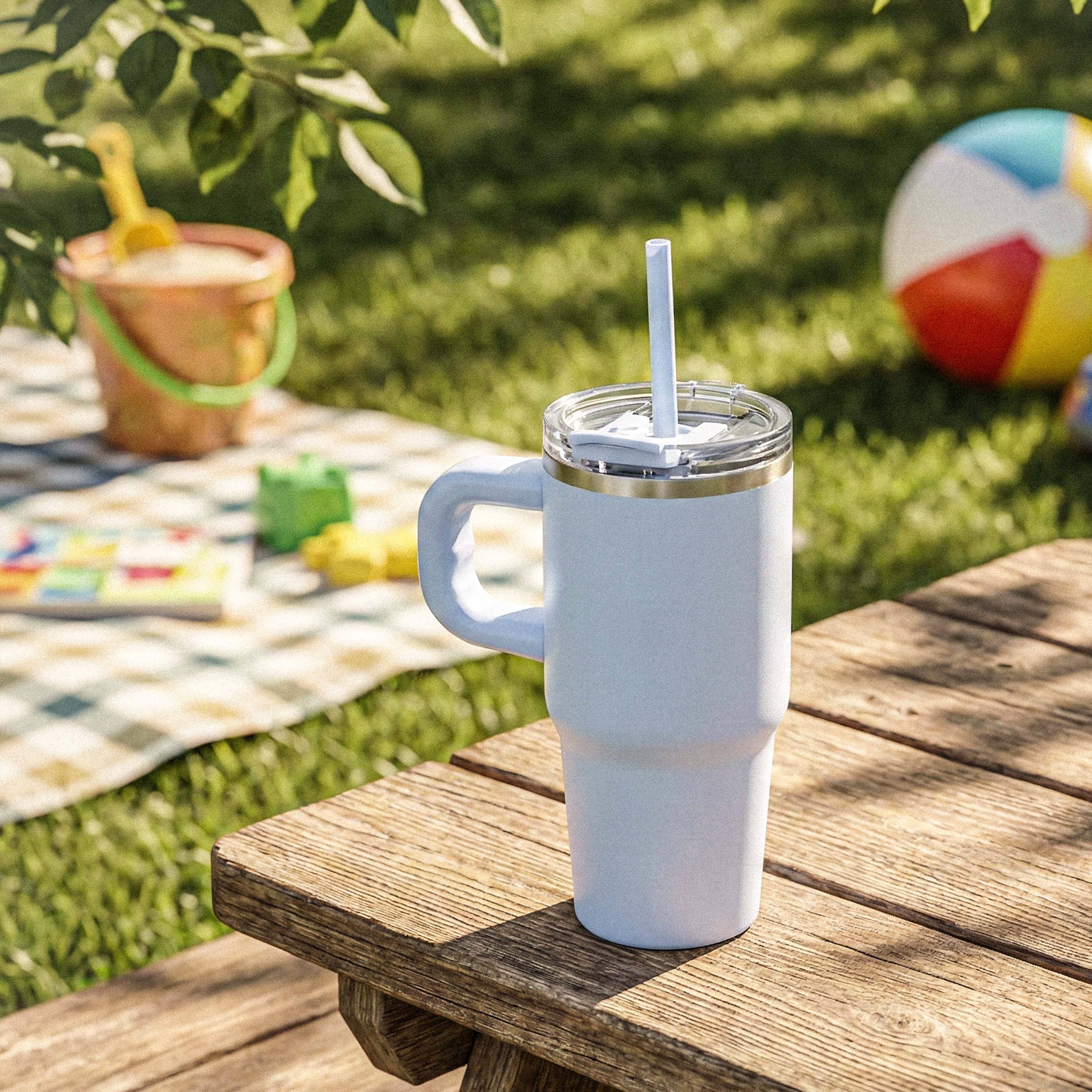 Insulated white tumbler with handle and straw on wooden picnic table outdoors, summer scene