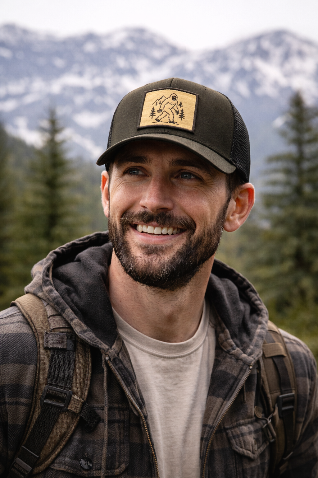 Bearded man in outdoor hiking gear and Sasquatch hat with mountains and trees in background