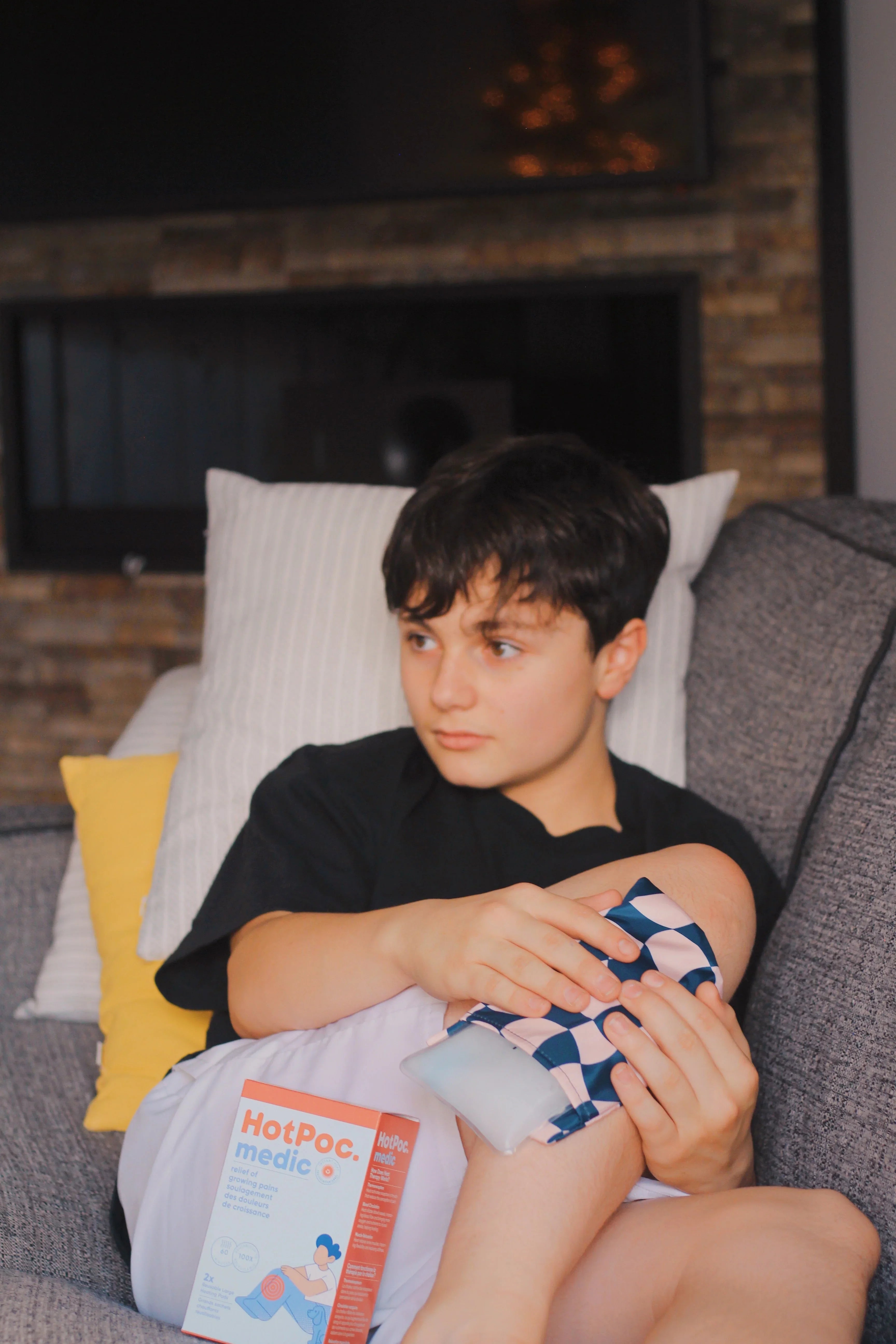 Boy using HotPac medic ice pack on knee for pain relief, sitting on couch at home