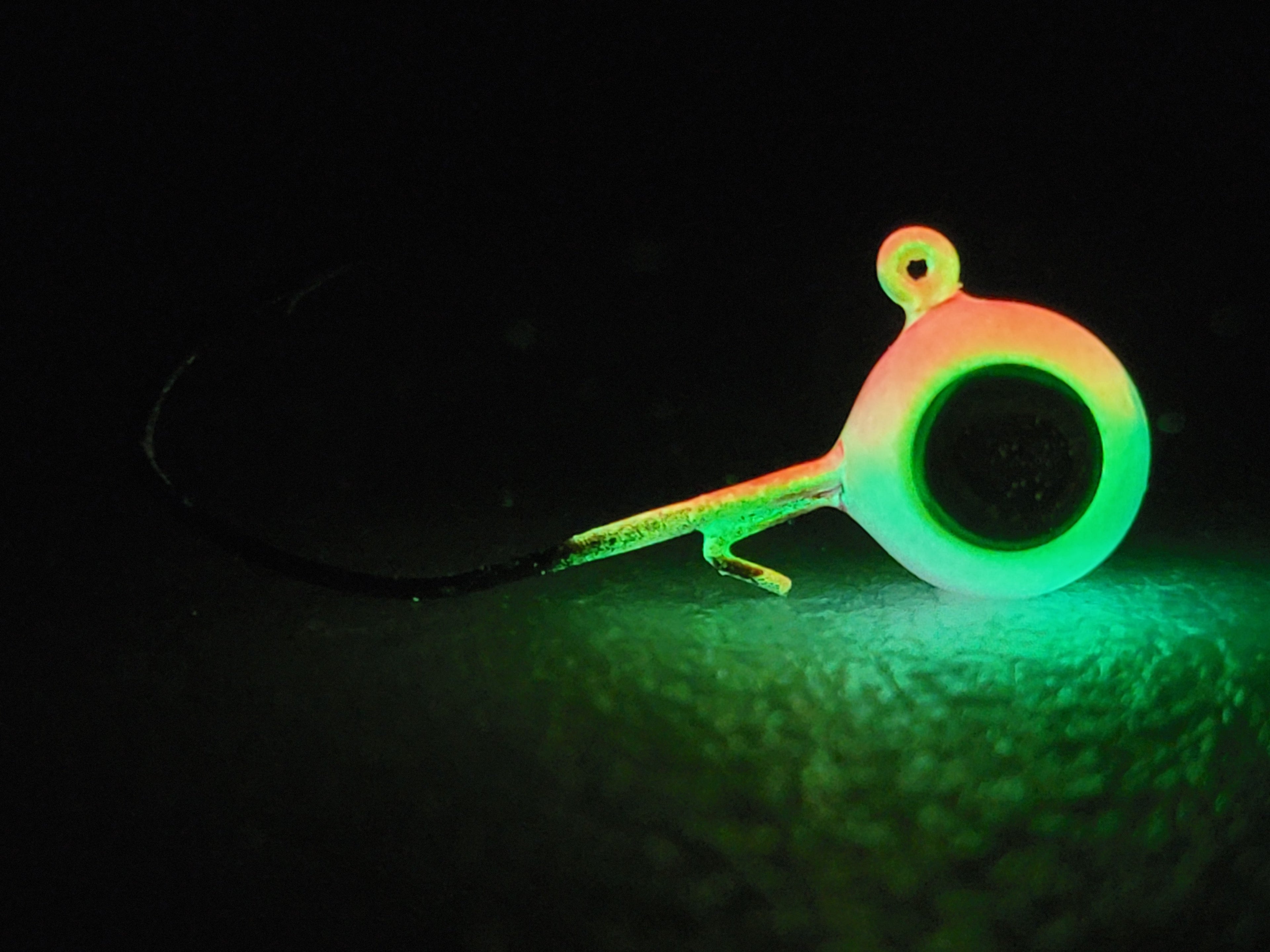 Glowing green and orange jig head fishing hook on dark textured surface, night fishing gear