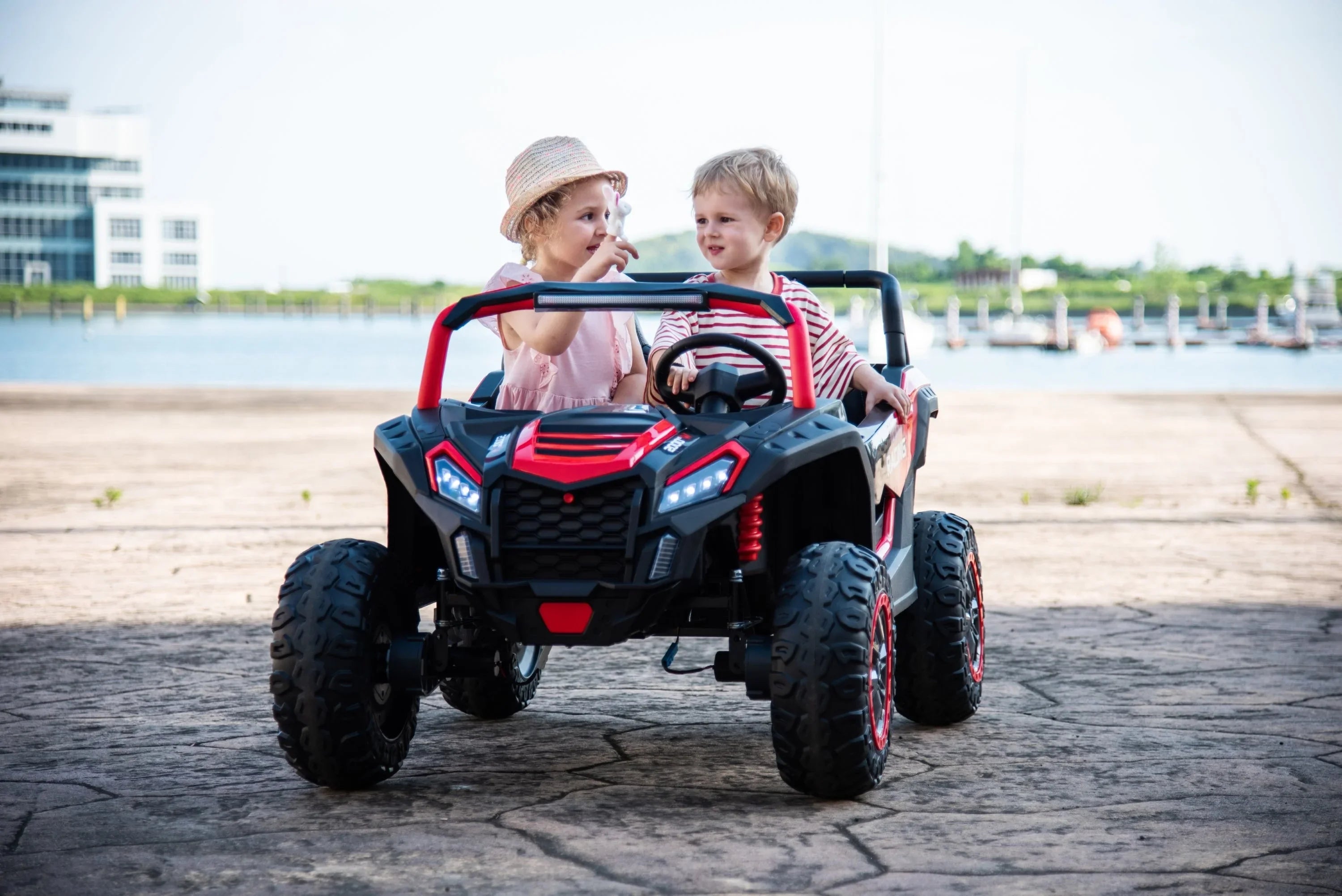 Two kids riding a black and red toy electric car outdoors near water