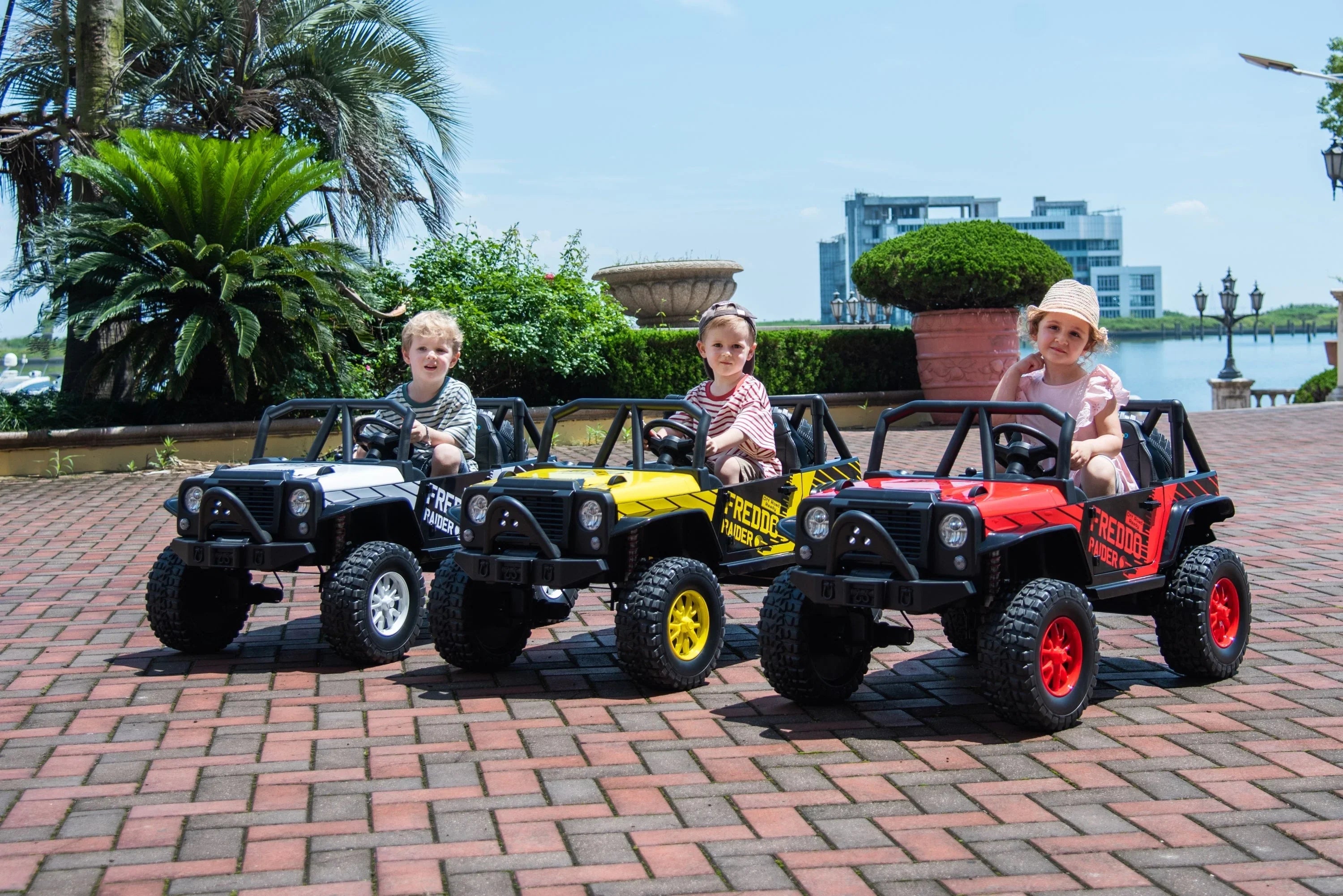 Kids driving toy electric cars outdoors on brick patio with greenery and modern buildings