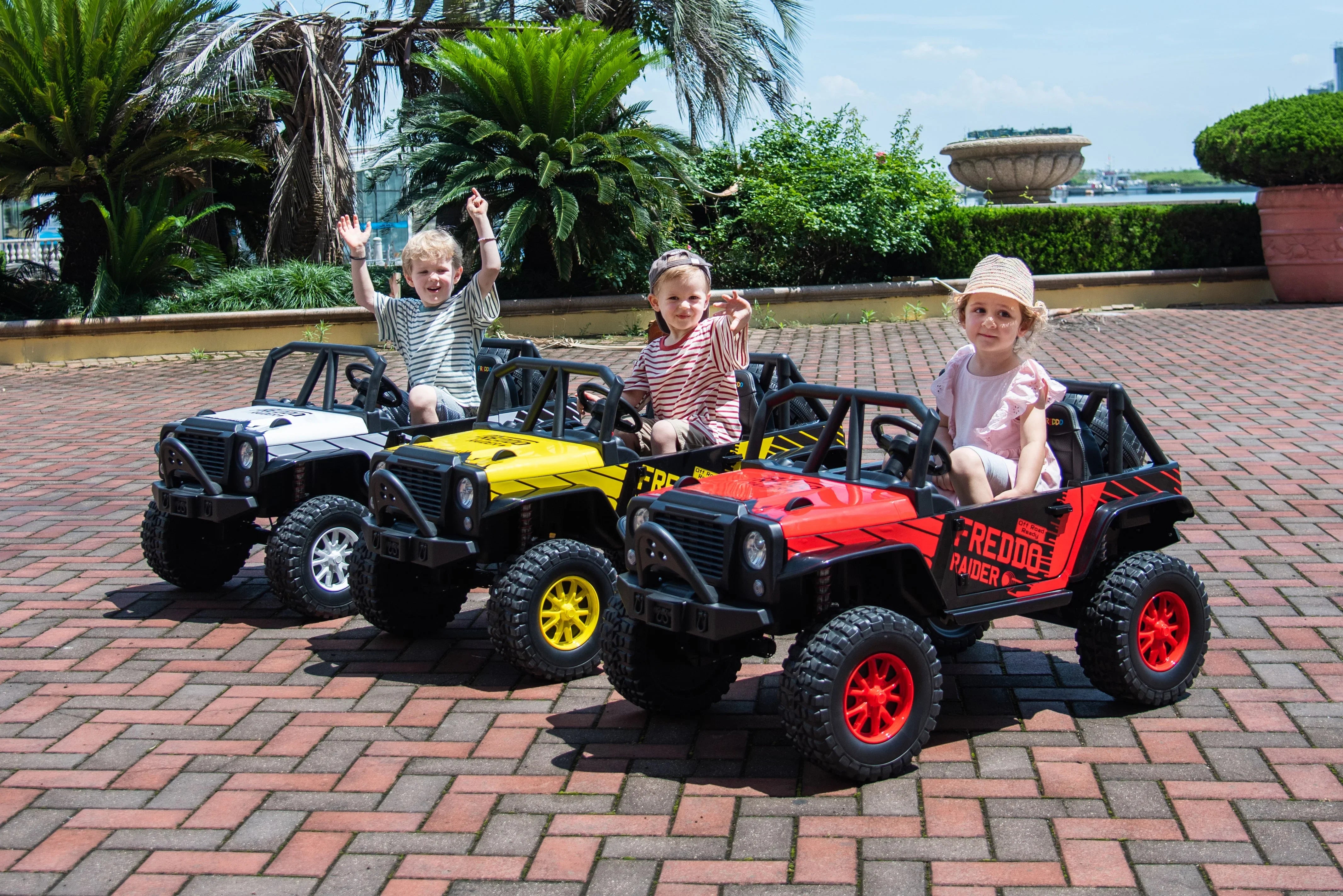 Three kids riding toy electric cars on brick patio with palm trees in background