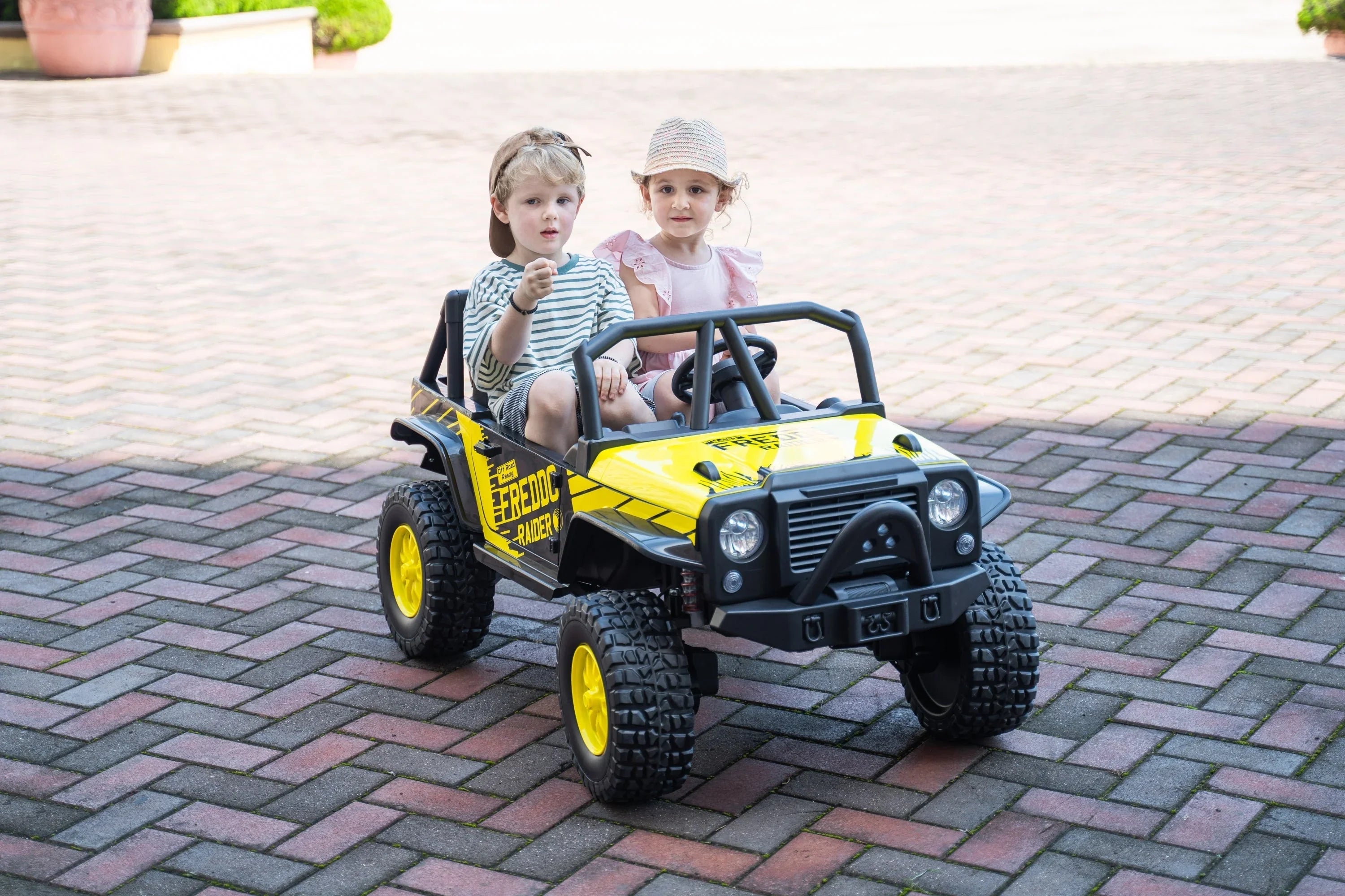 Two kids riding a yellow and black electric toy jeep on a paved driveway