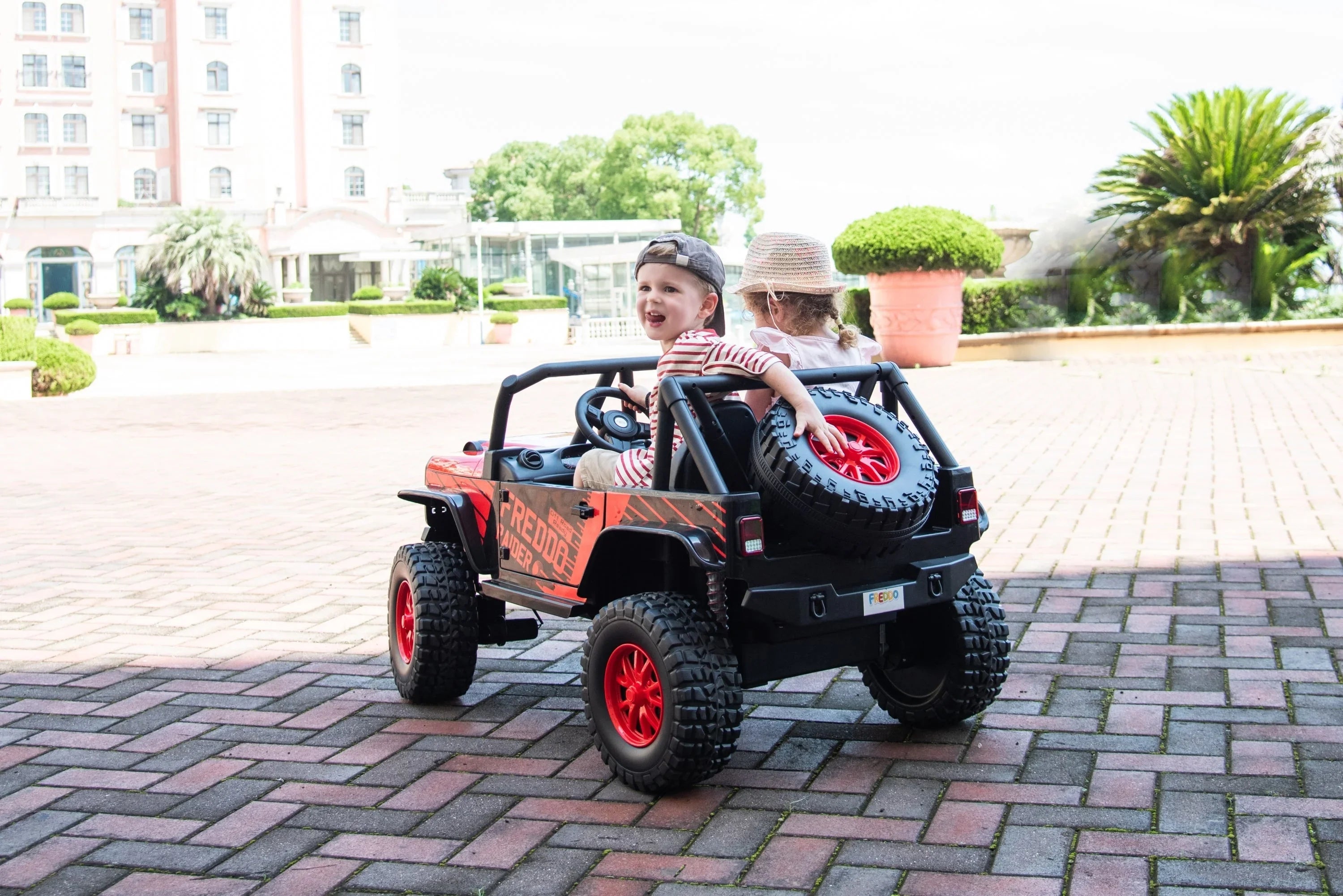 Two kids riding a Freddo Raider electric toy car outdoors on a sunny brick patio