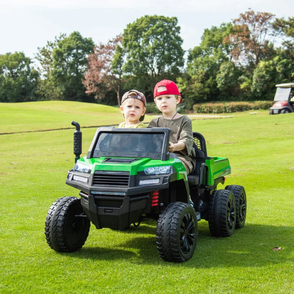 Two kids riding a green electric toy truck outdoors on a grassy field
