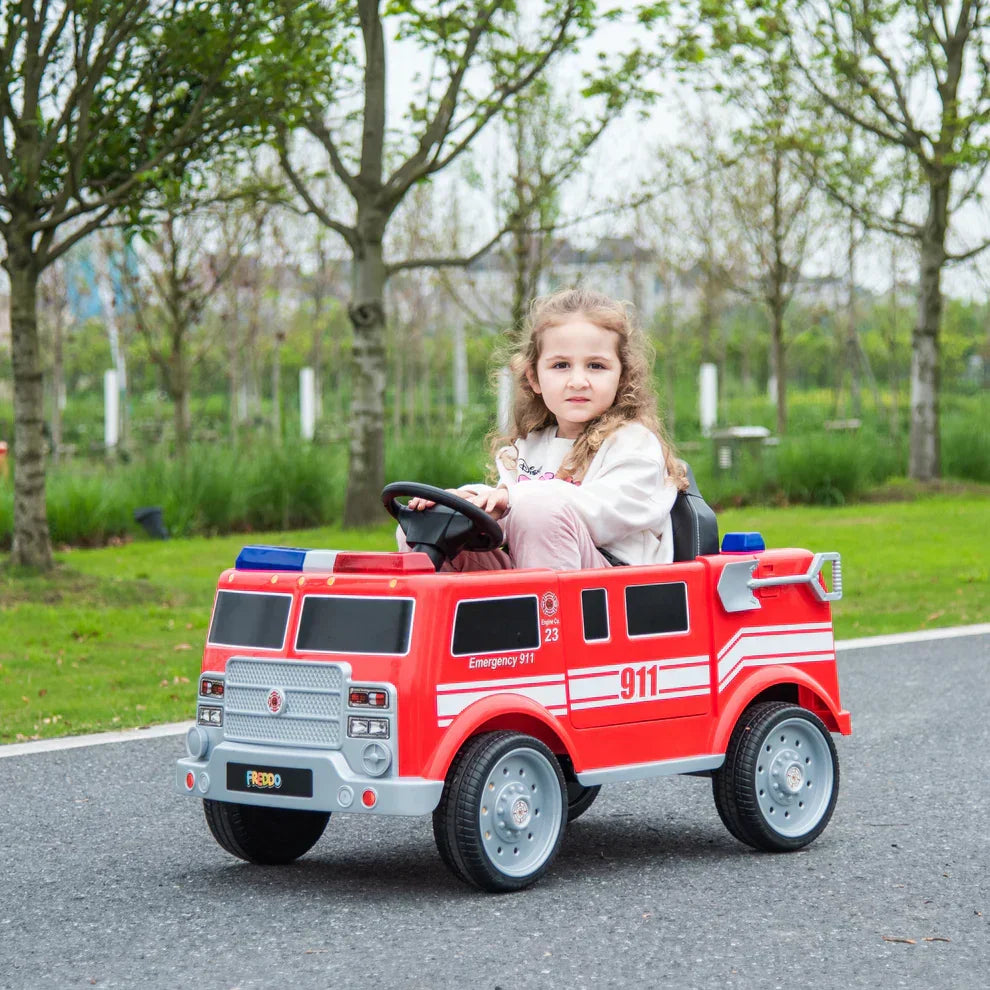 Girl riding red toy fire truck car outdoors on park path with trees in background