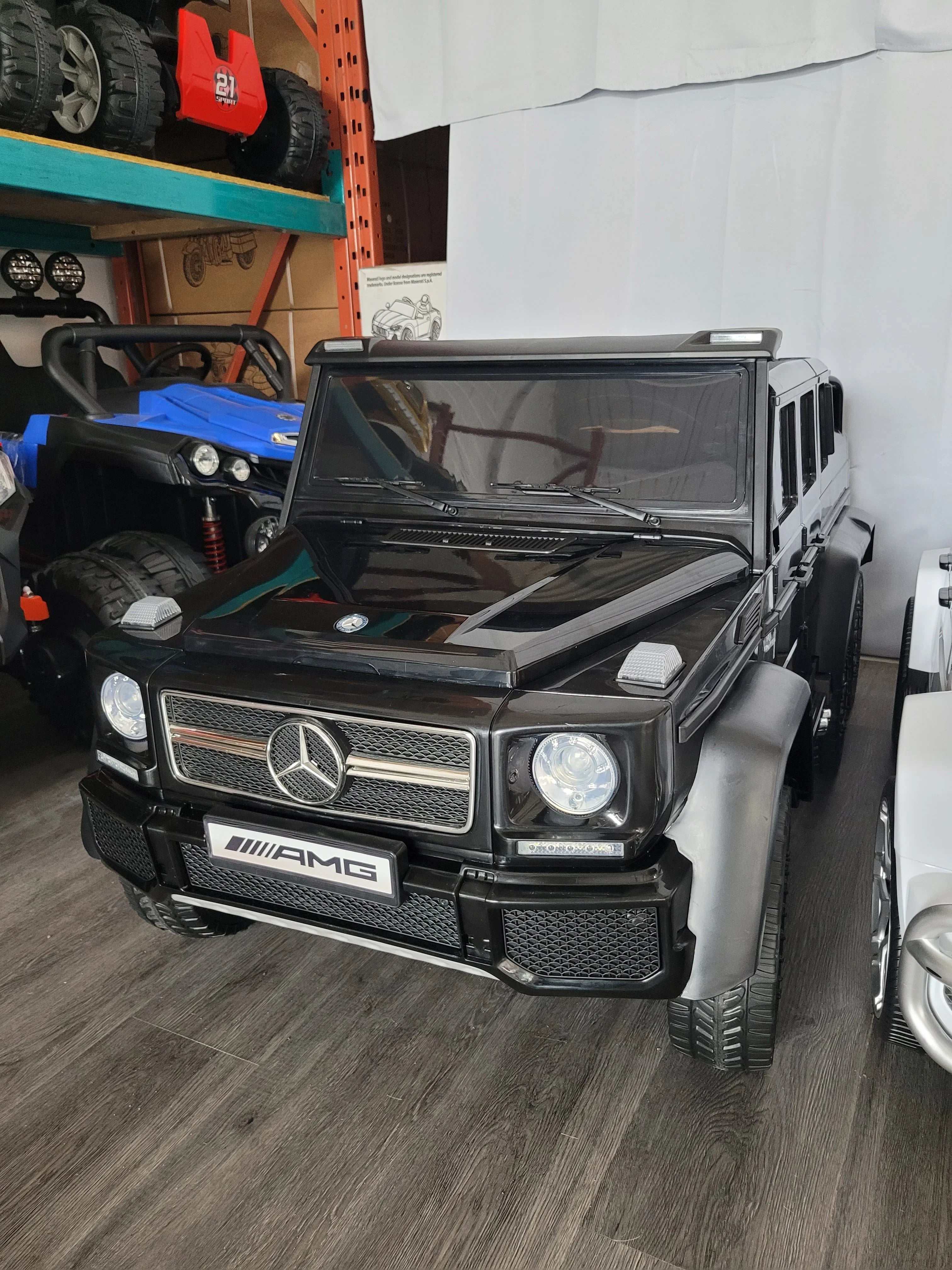Black Mercedes AMG G-Class kids electric car on display indoors with toy vehicles in background.