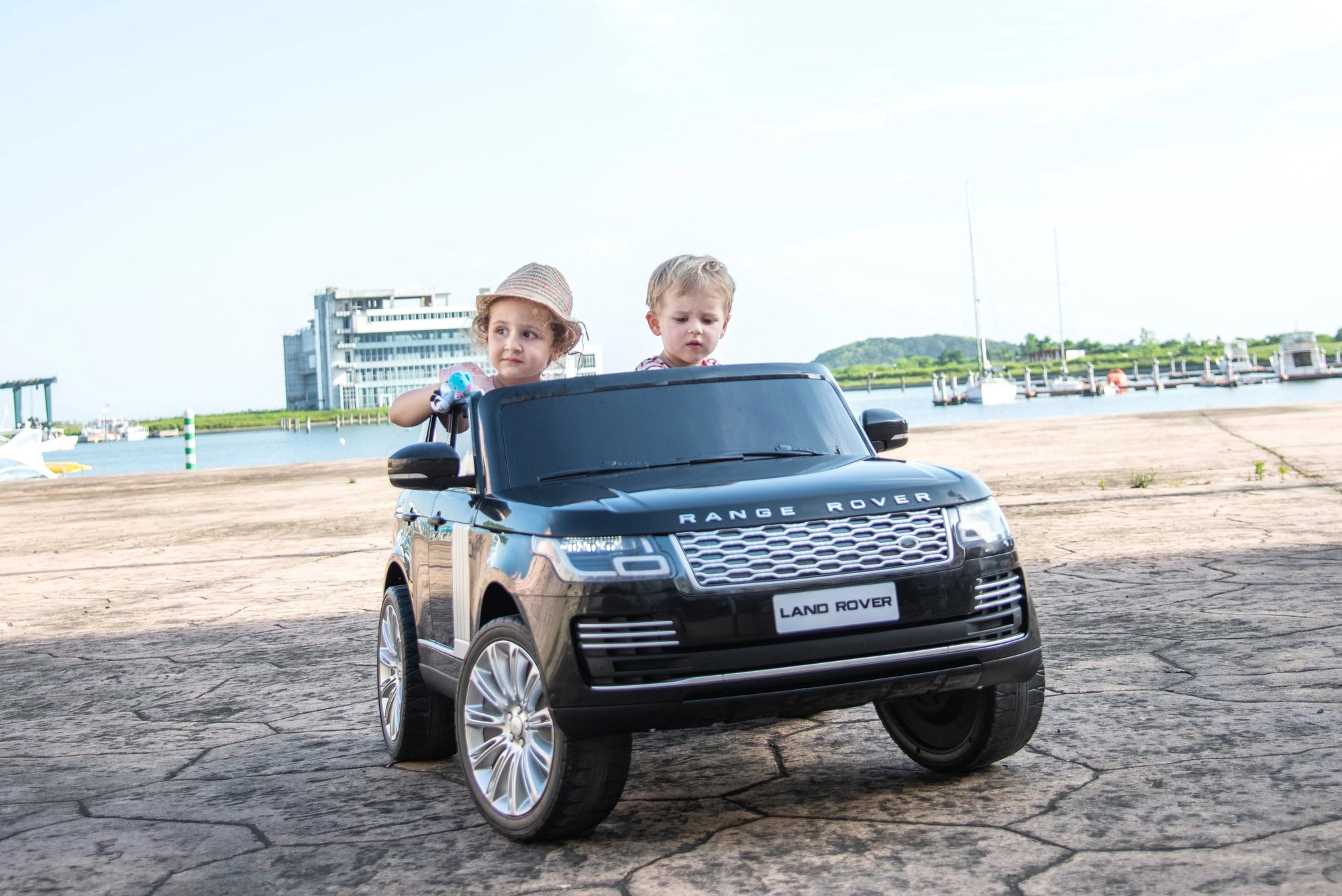 Two kids in toy Range Rover car outdoors near marina, waterfront, and boats