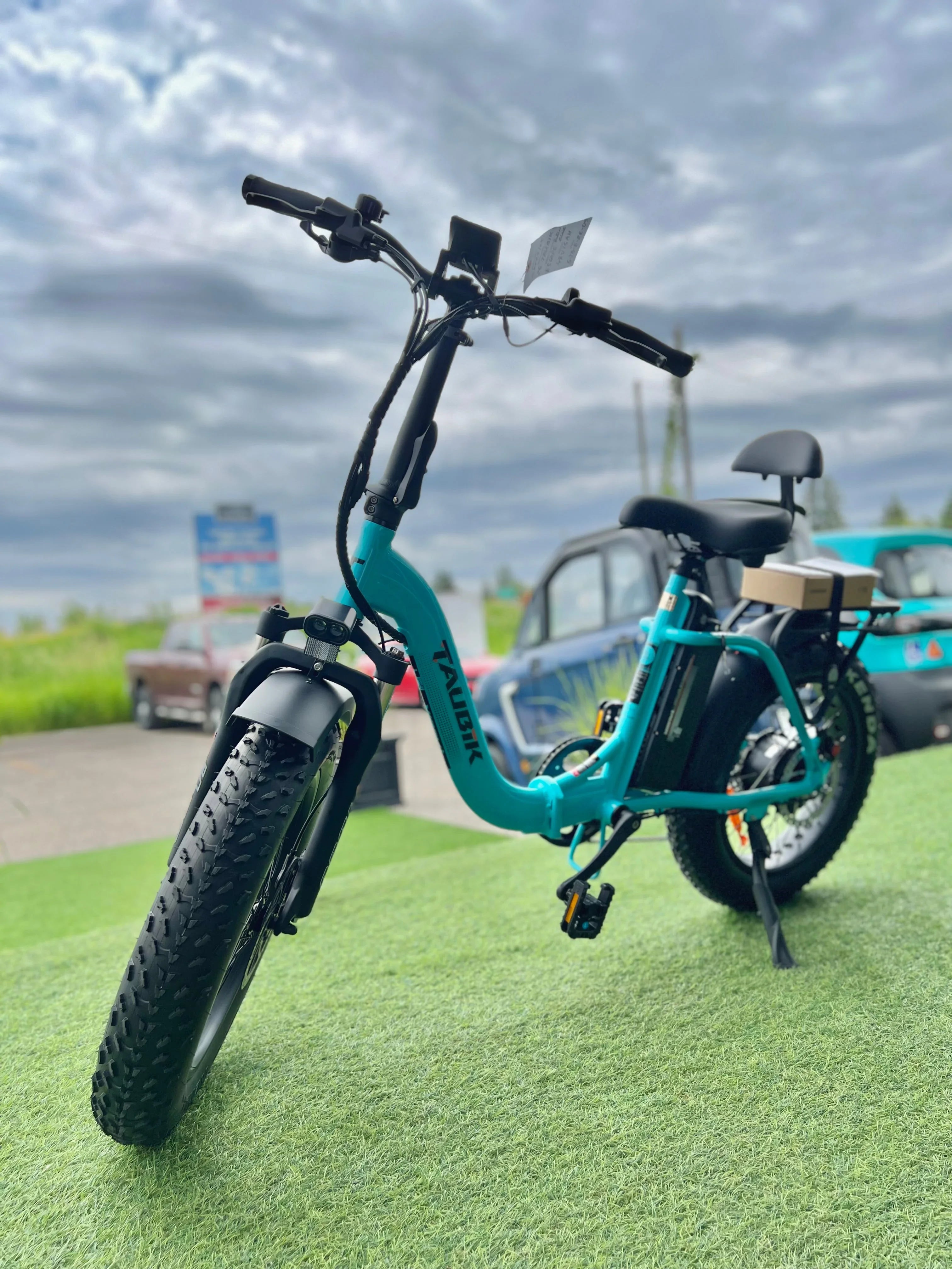 Teal TAUBIK electric bike displayed on grass with cars and cloudy sky in background