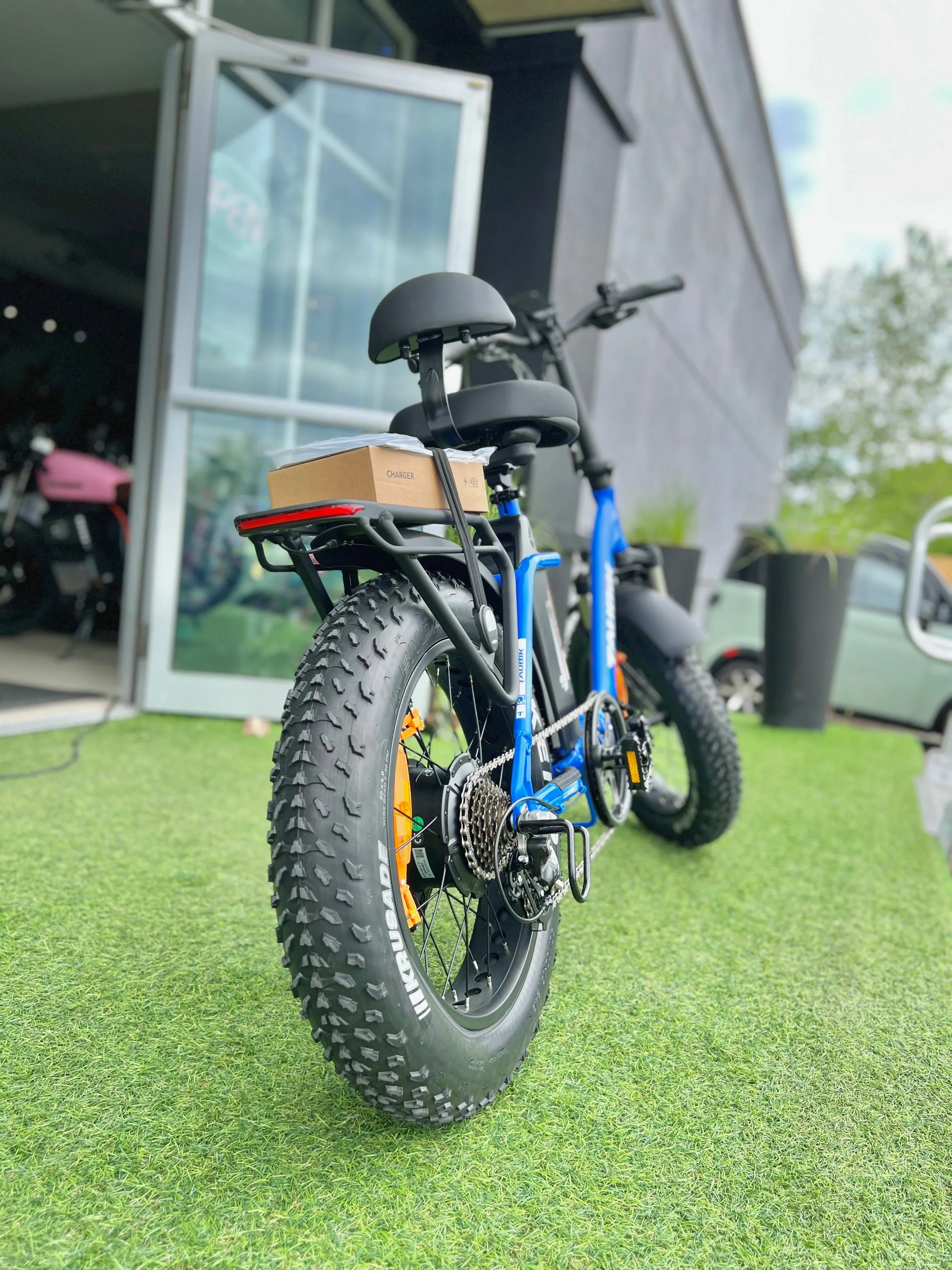 Blue fat tire electric bike with rear rack and box, parked on artificial grass outside store