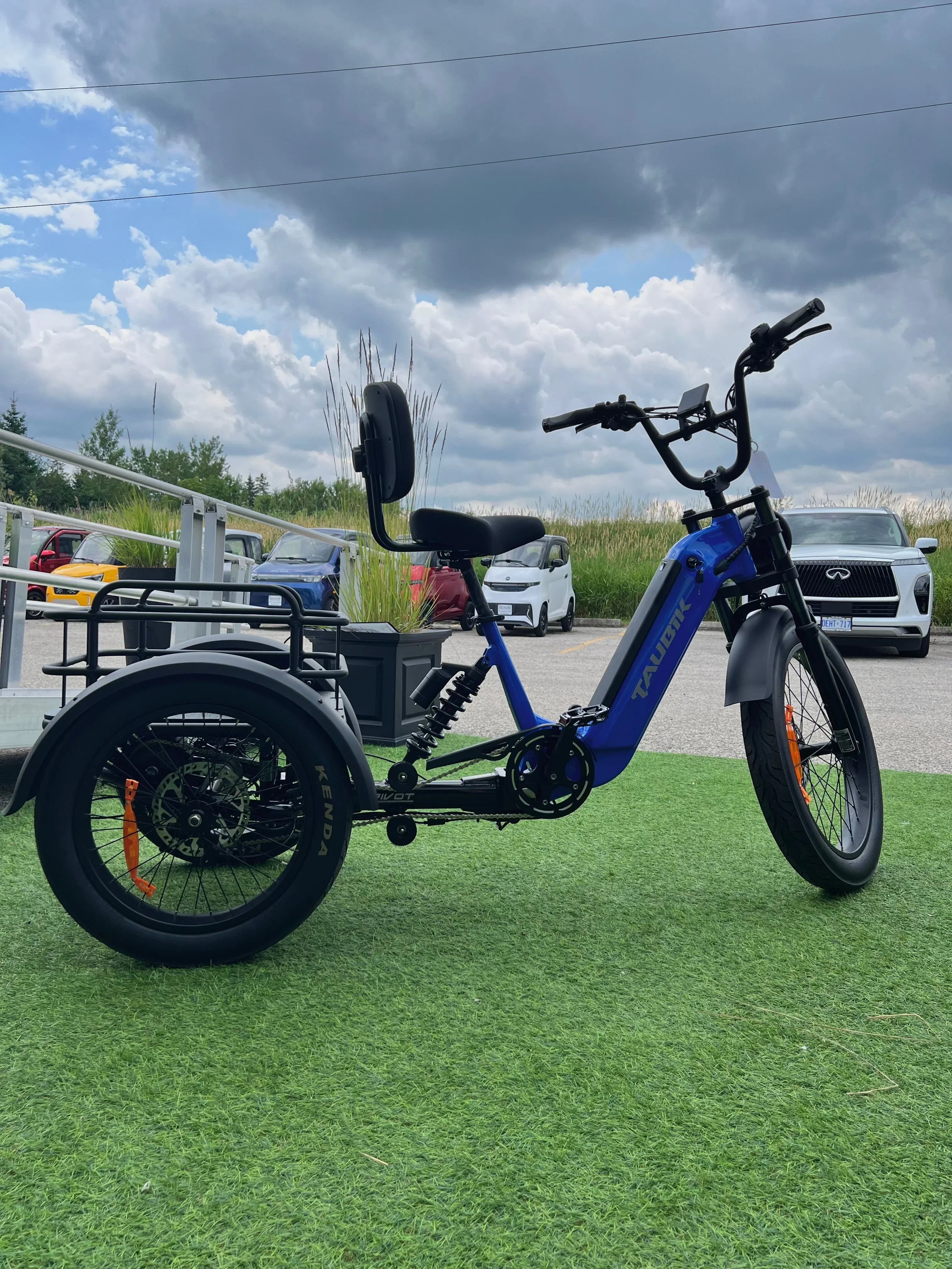 Blue electric trike with backrest, on green turf, parked outdoors with cars in background