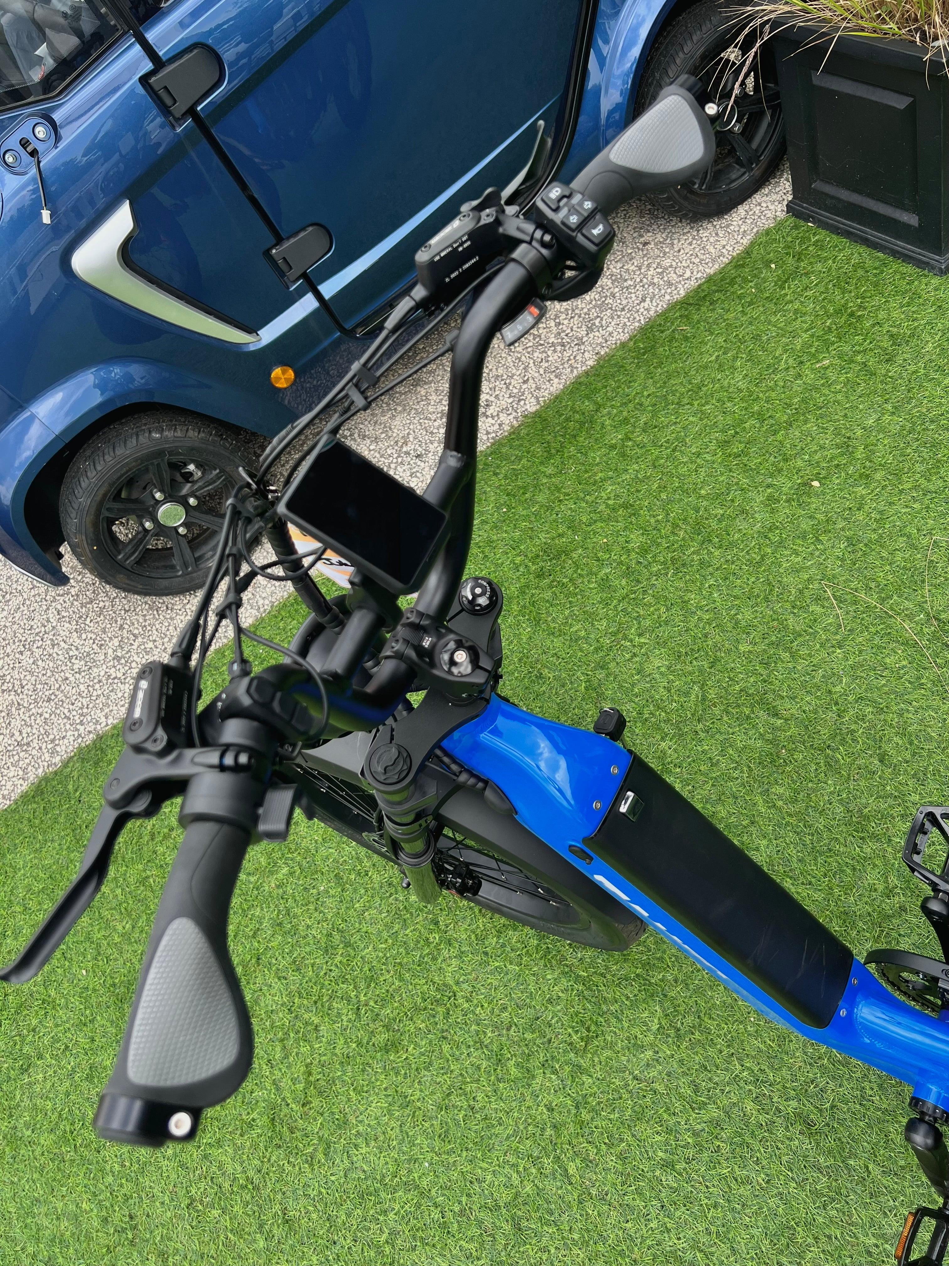 Blue electric bike handlebar with display, parked on artificial grass beside a compact blue car