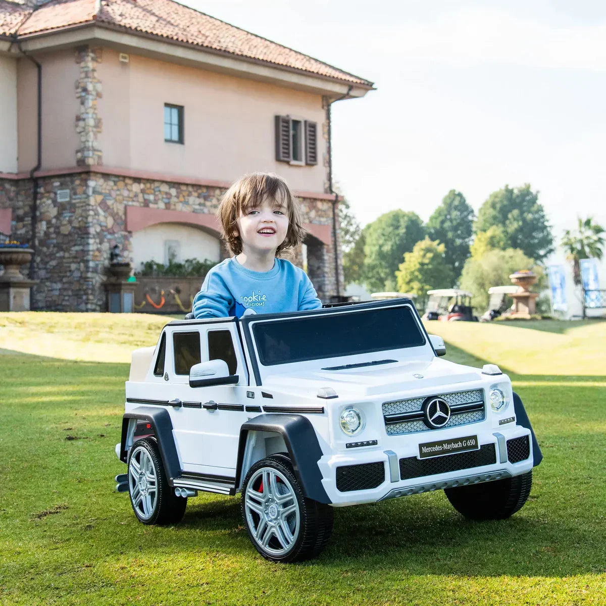 Smiling child in a white Mercedes-Maybach G650 kids ride-on car outdoors on grass