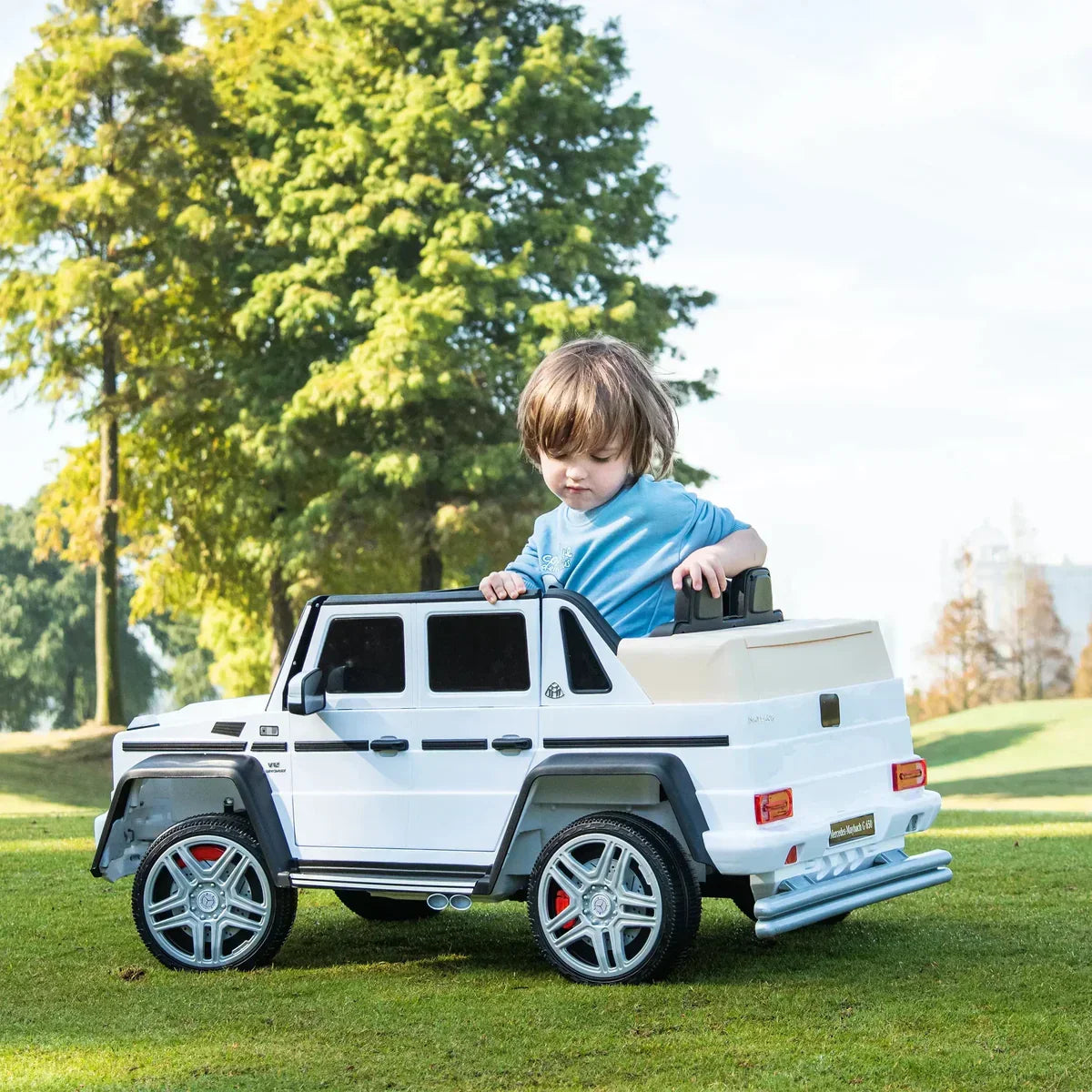 Child in a white ride-on toy SUV car on green grass with trees in background outdoors