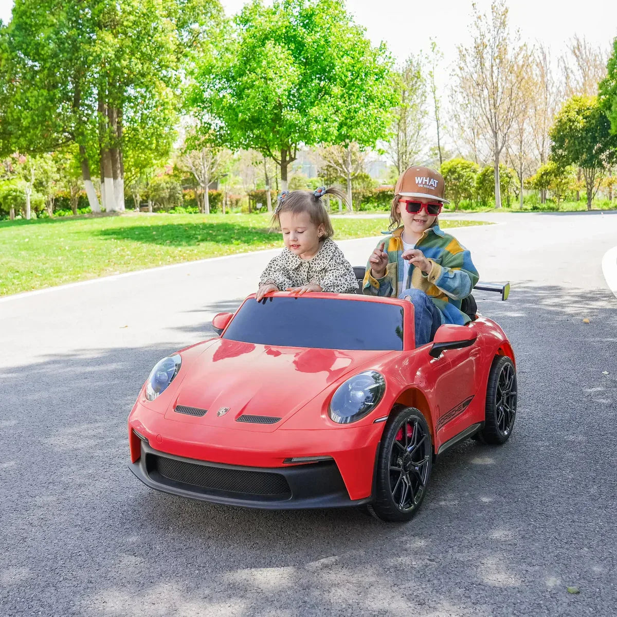 Two kids riding a red Porsche electric toy car outdoors on a sunny day