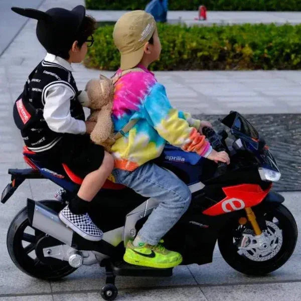 Two kids riding blue electric motorcycle bike outdoors, one holding a teddy bear.