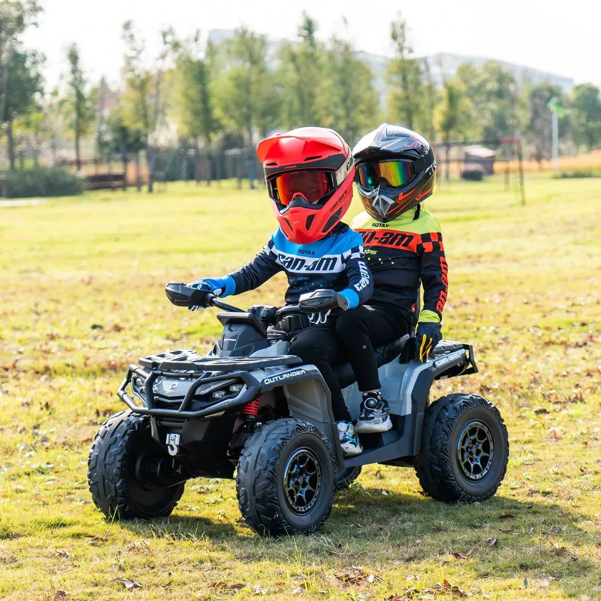 Two kids riding Can-Am Outlander 24V ride-on ATV toy outdoors on grass field