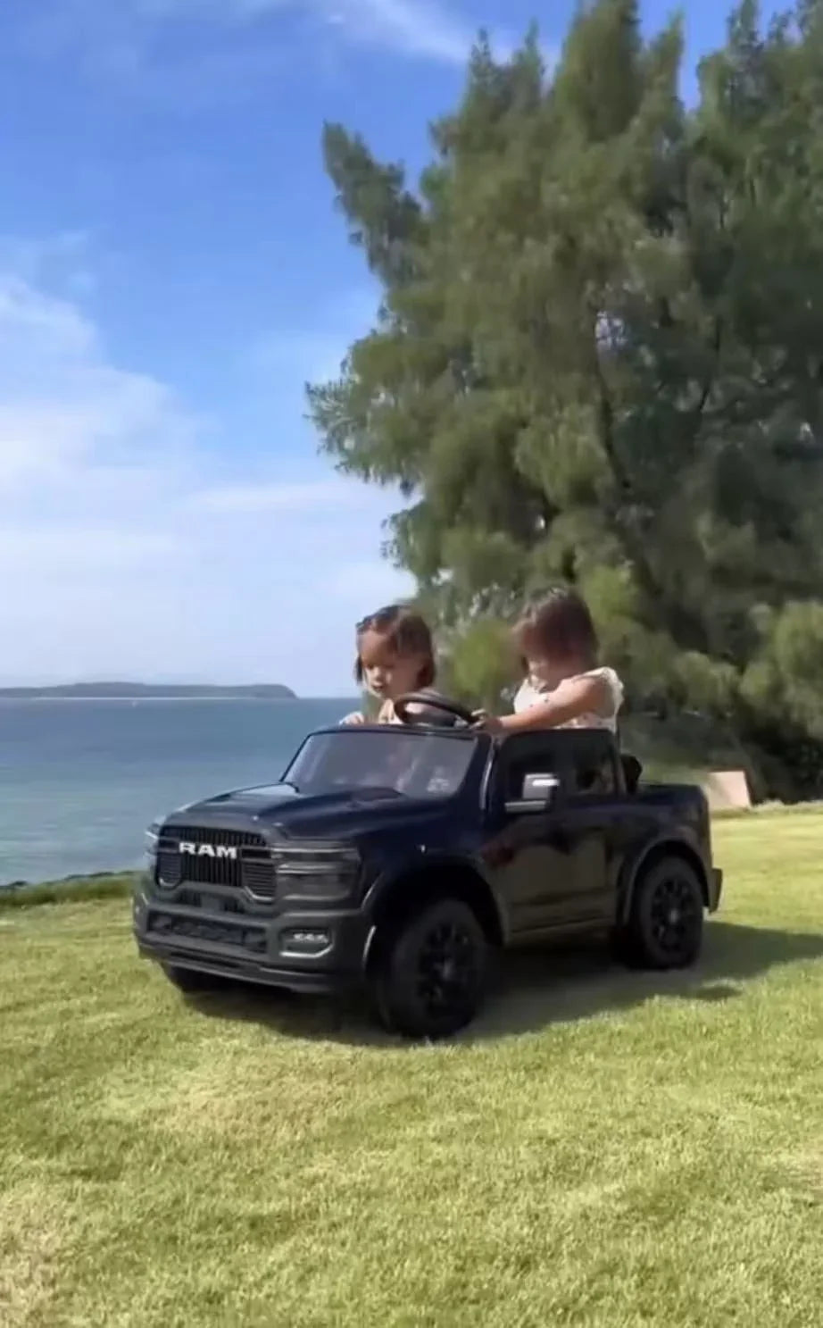 Two kids in a black RAM toy truck on grass near the beach with trees and blue sky