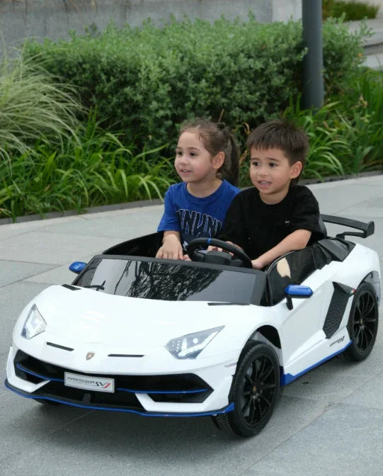 Two kids riding a white toy Lamborghini car outdoors on a paved path