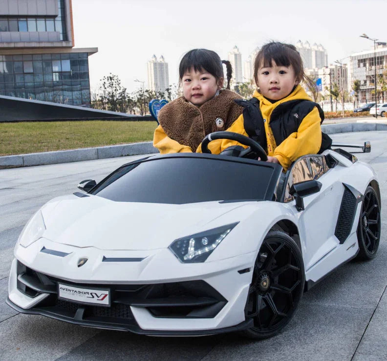 Two children in a white toy Lamborghini Aventador SV car outdoors in a city park