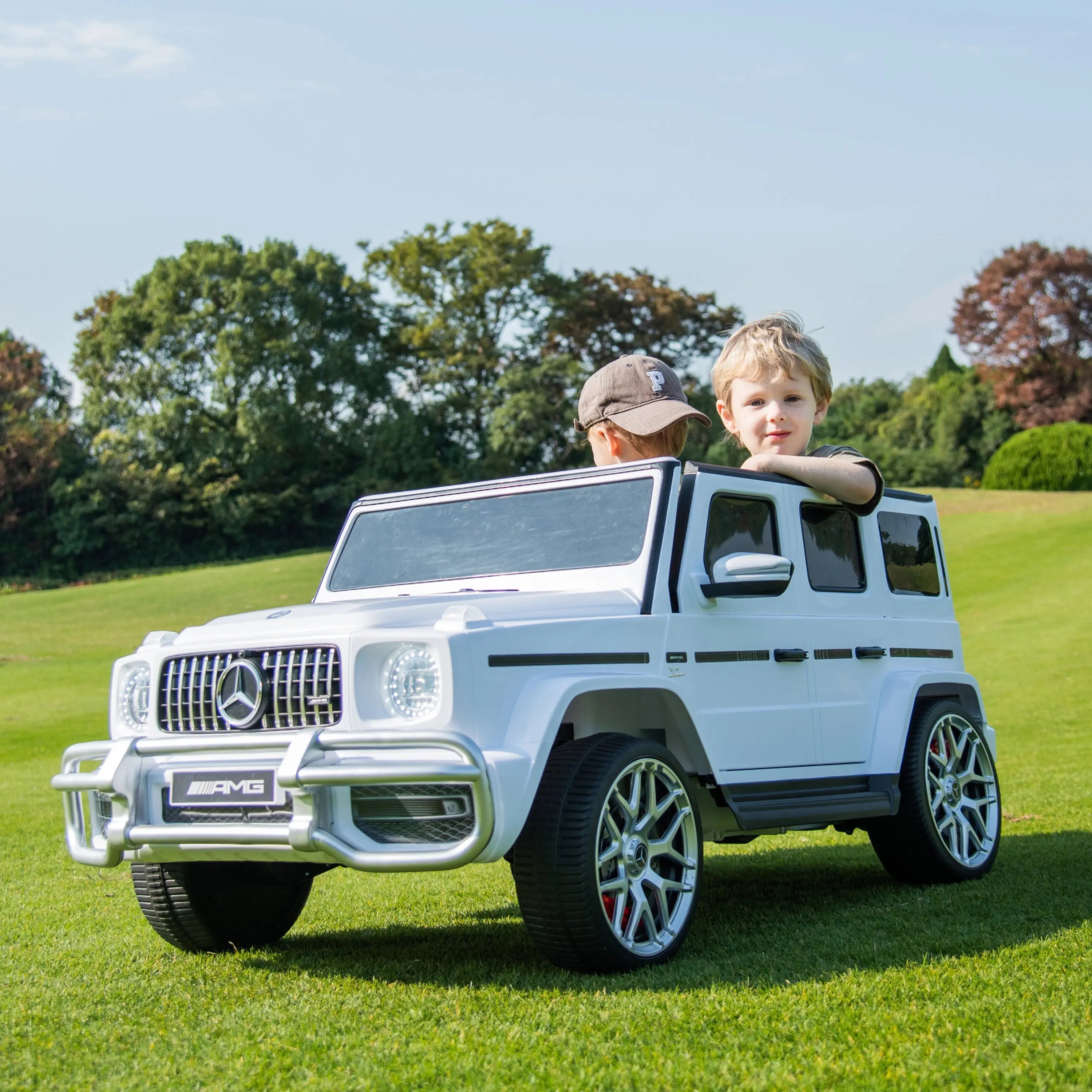 Two kids in white Mercedes-Benz G63 AMG ride-on toy car outdoors on grass