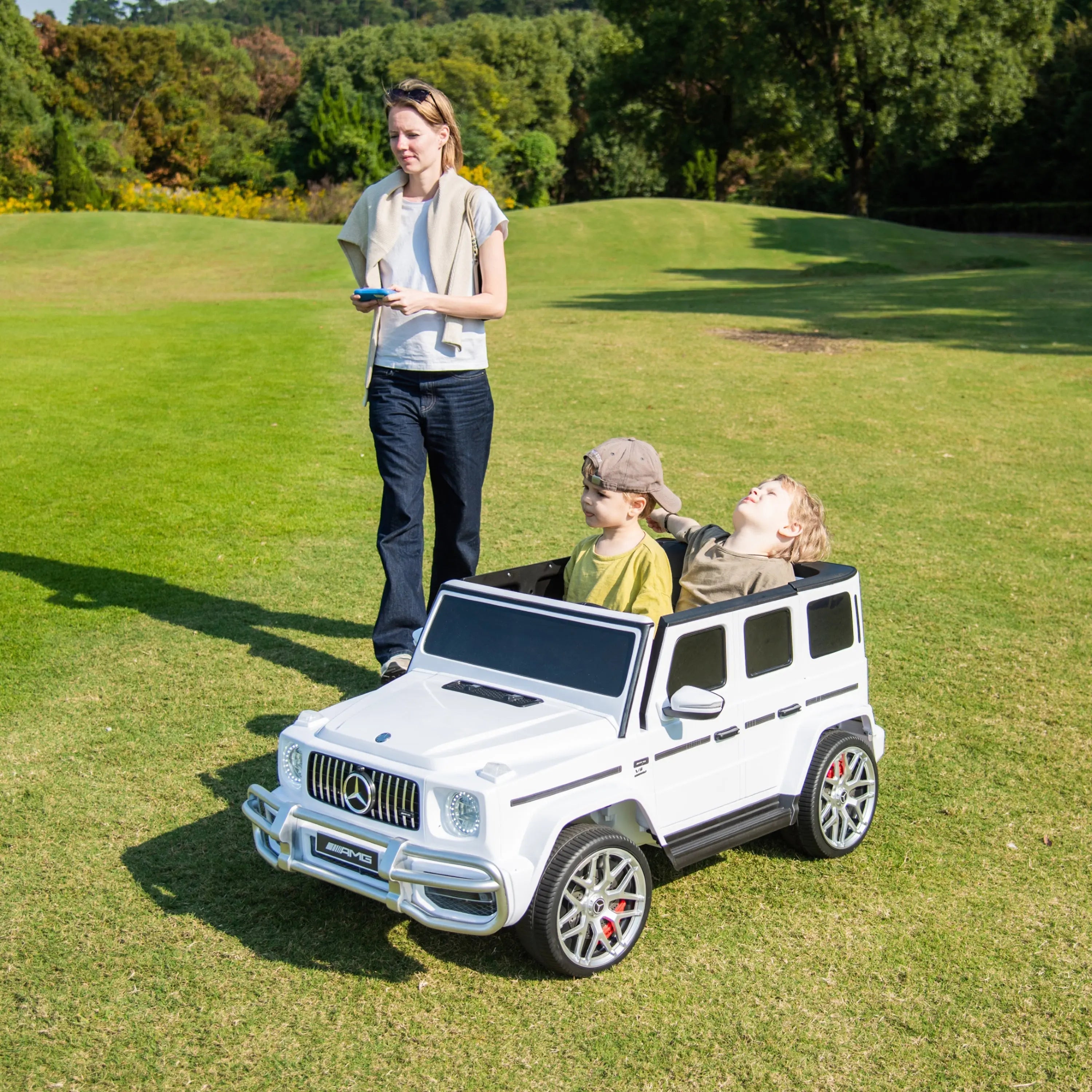 Two kids riding in a white Mercedes-Benz G63 AMG toy car on grass with adult supervising