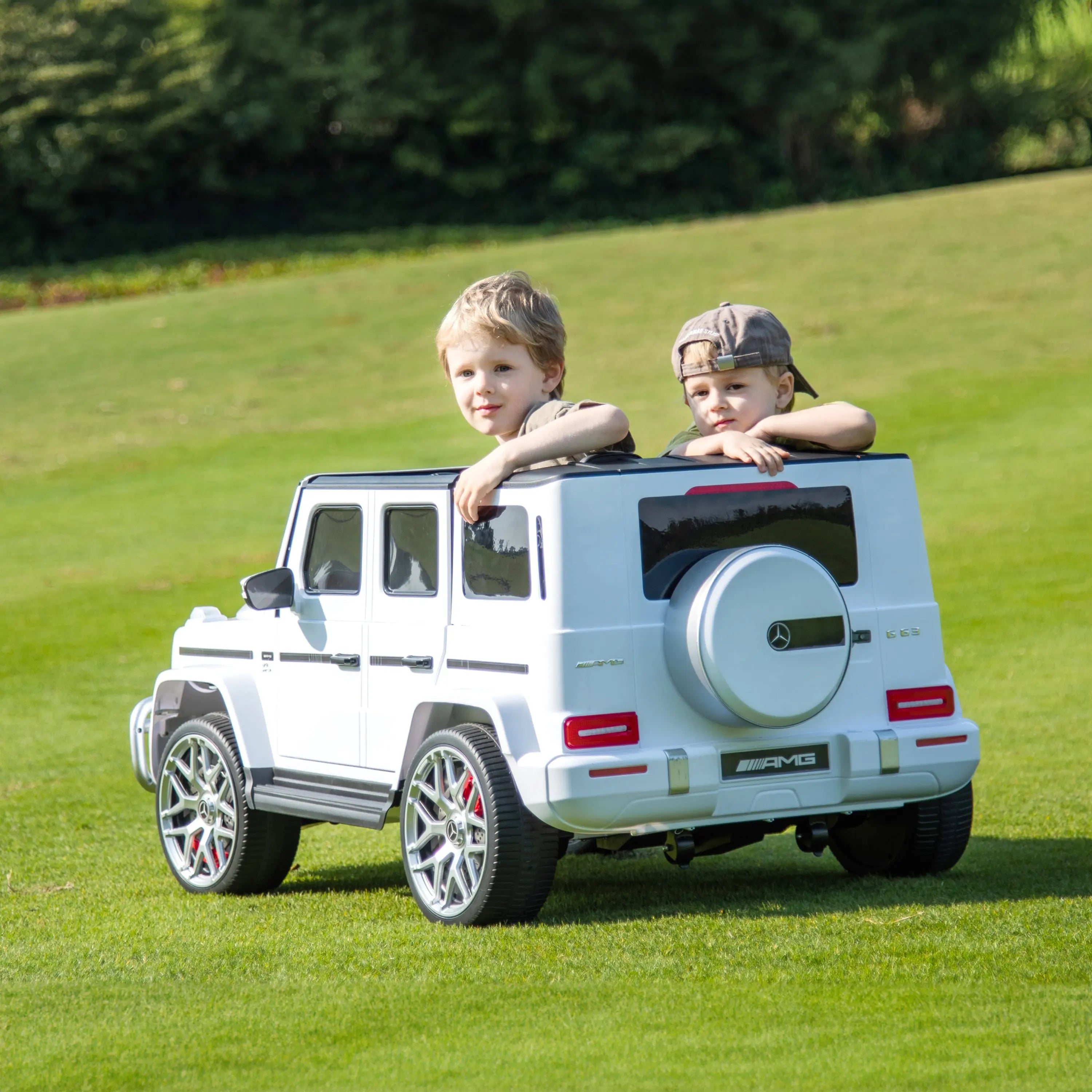 Two kids in a white 24V Mercedes G63 AMG ride-on car on green grass outdoors