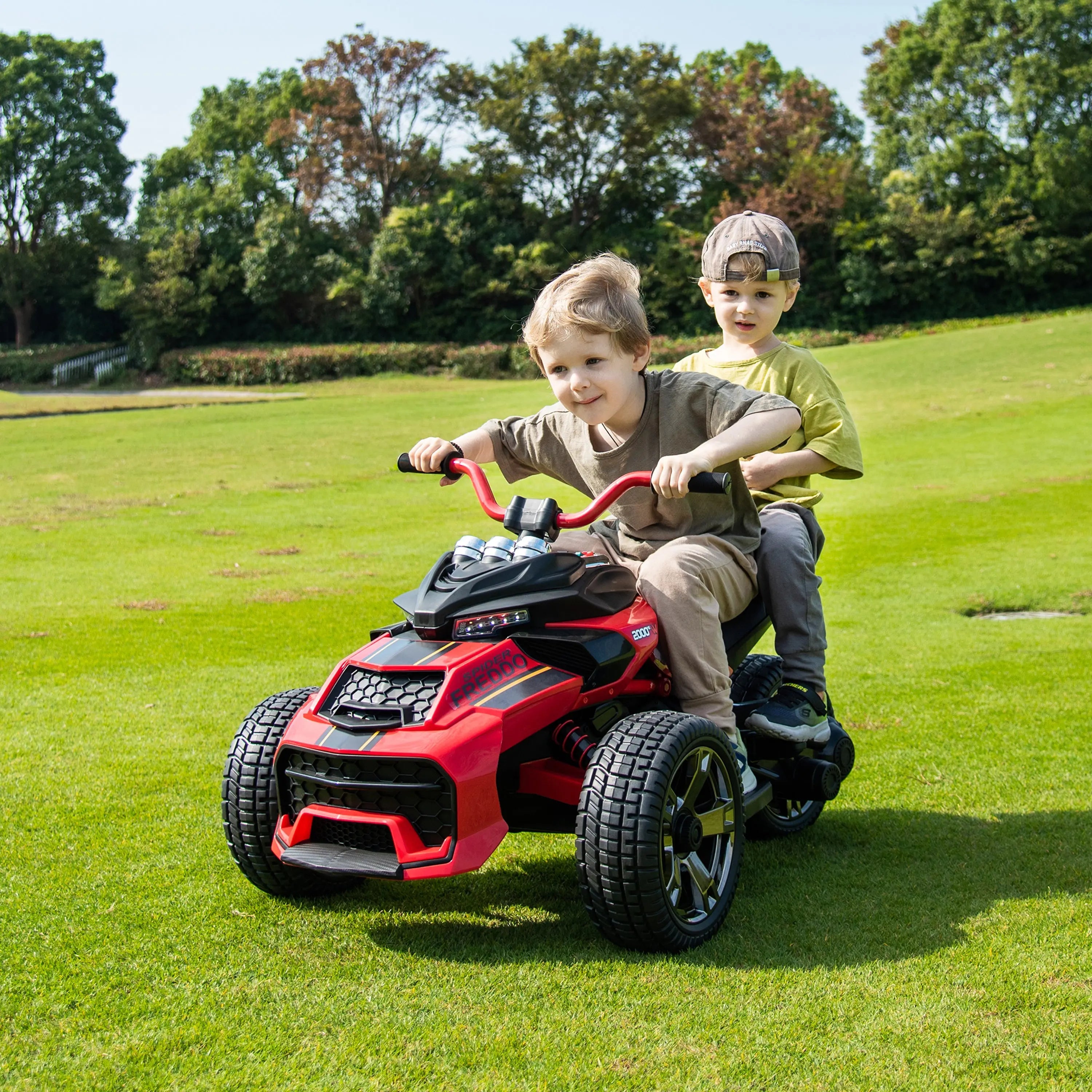 Two children riding a red 24V three-wheel ride-on motorcycle on grass outdoors