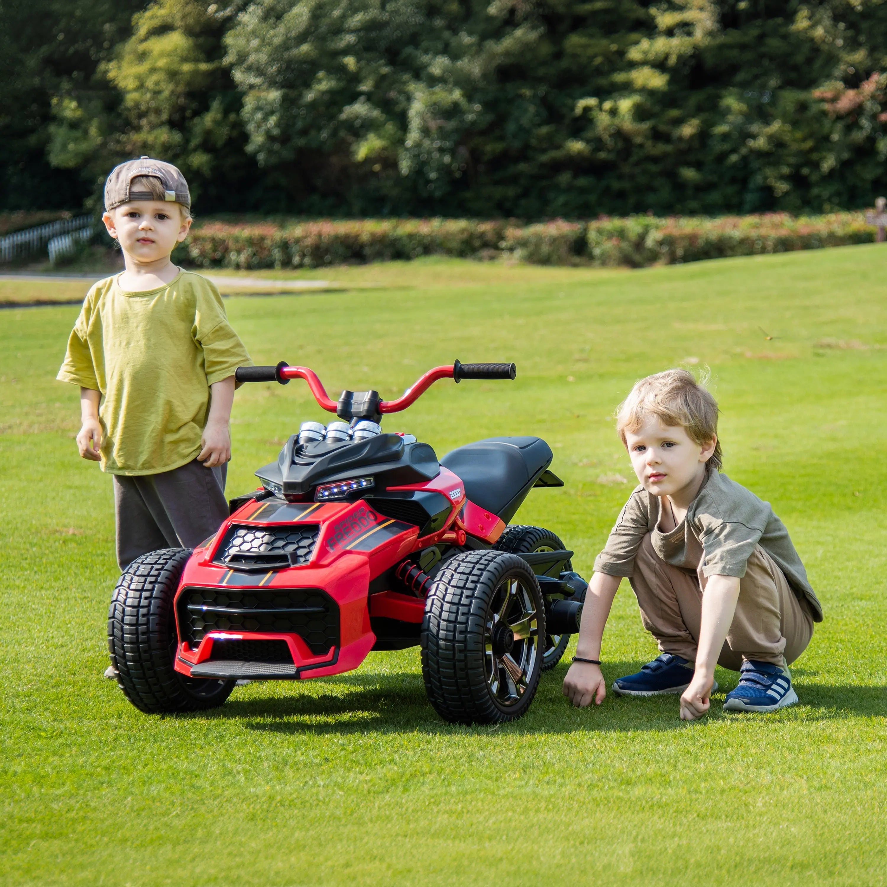 Two kids with a red 24V three-wheel ride-on motorcycle outdoors on grass
