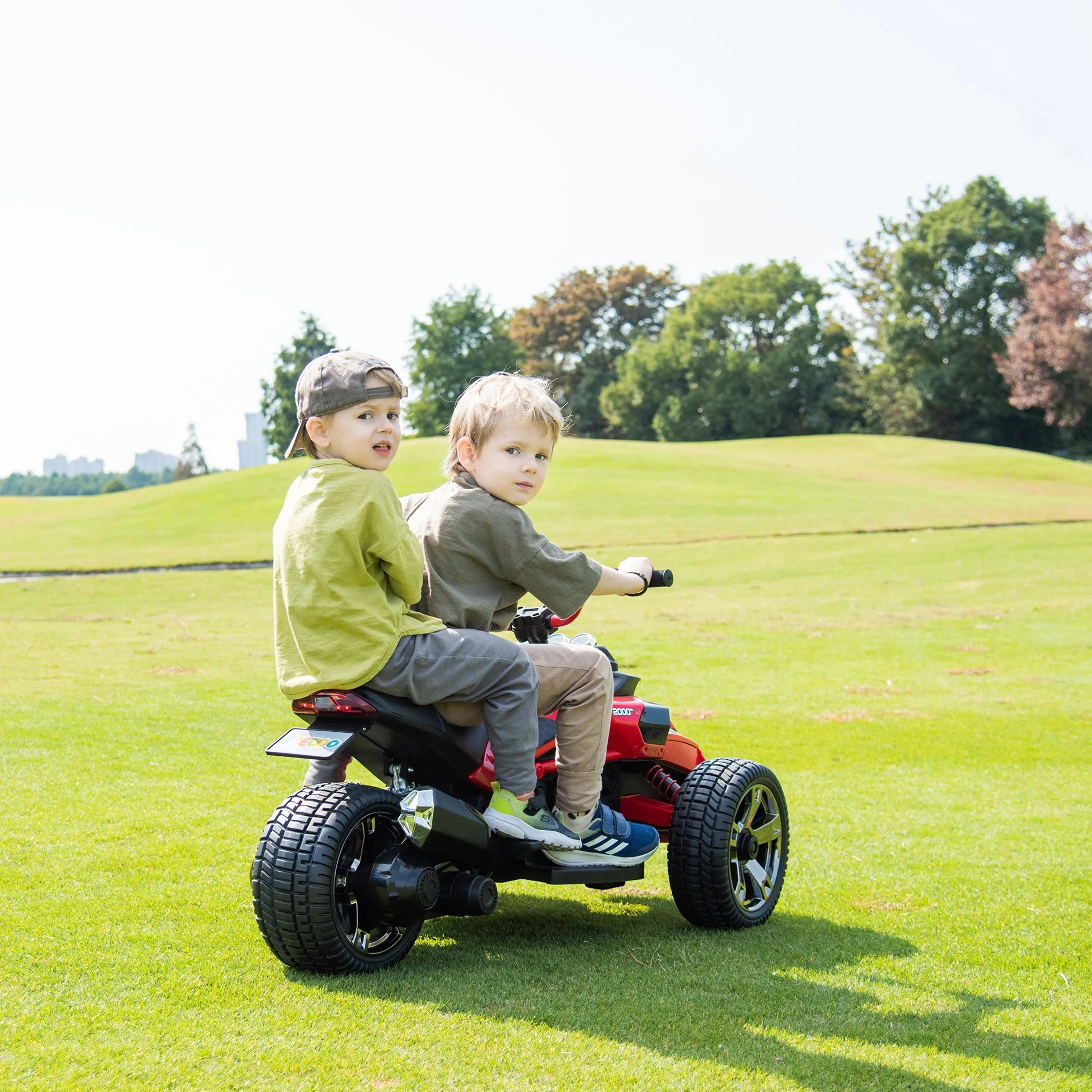 Two kids riding a red 3-wheel electric motorcycle outdoors on grass