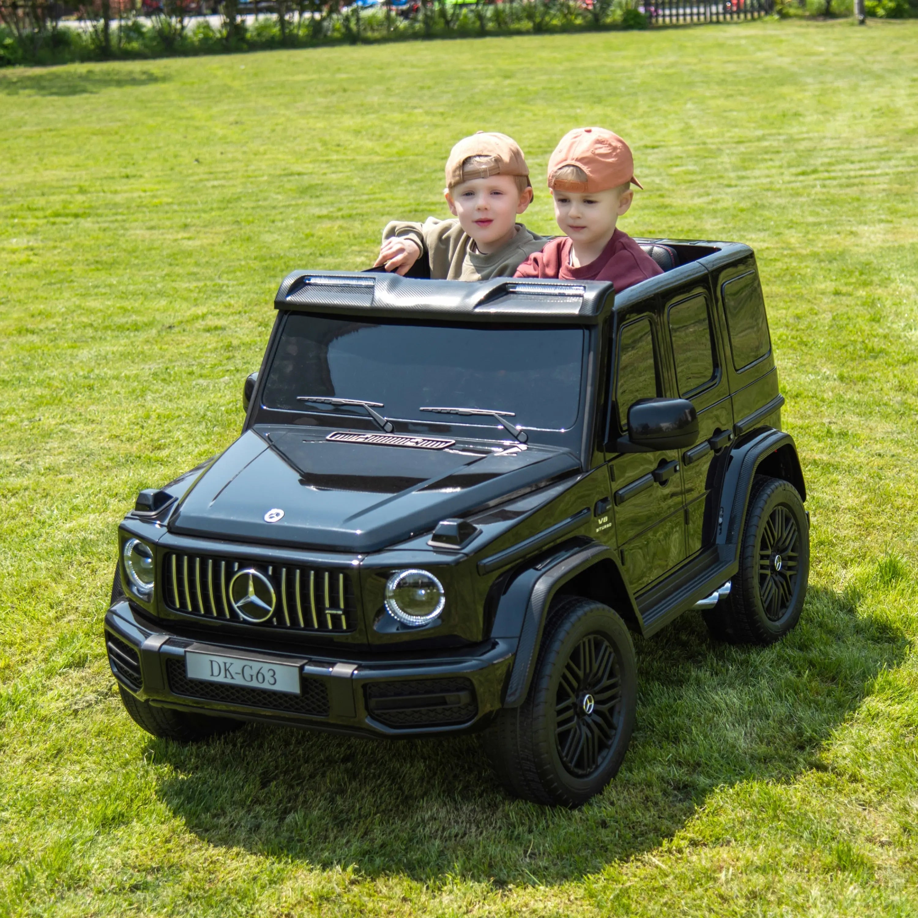 Two children riding black 24V Mercedes G-Wagon toy car on grass outdoors