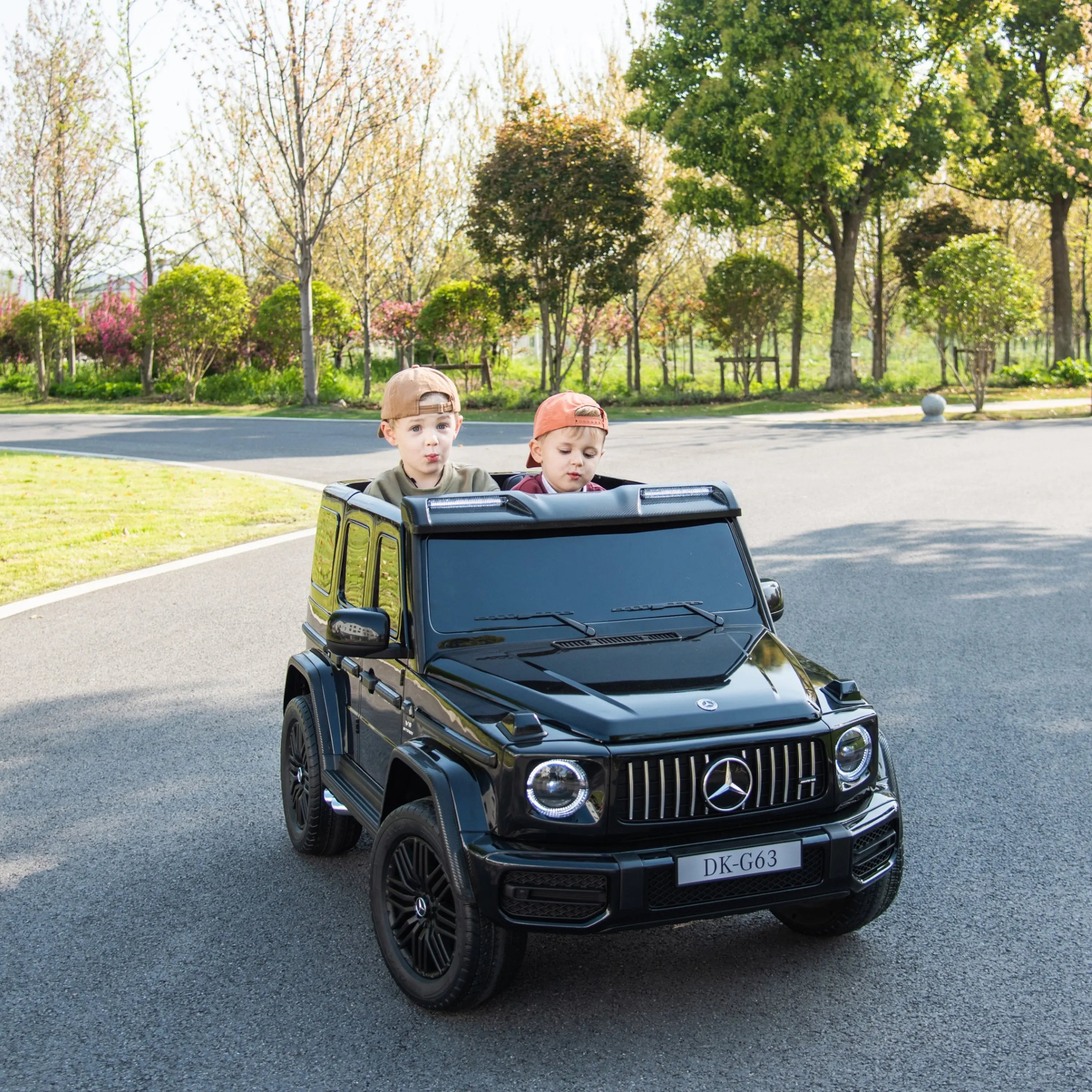 Two kids ride a black 24V Mercedes-Benz G-Wagon toy car outdoors on a sunny day