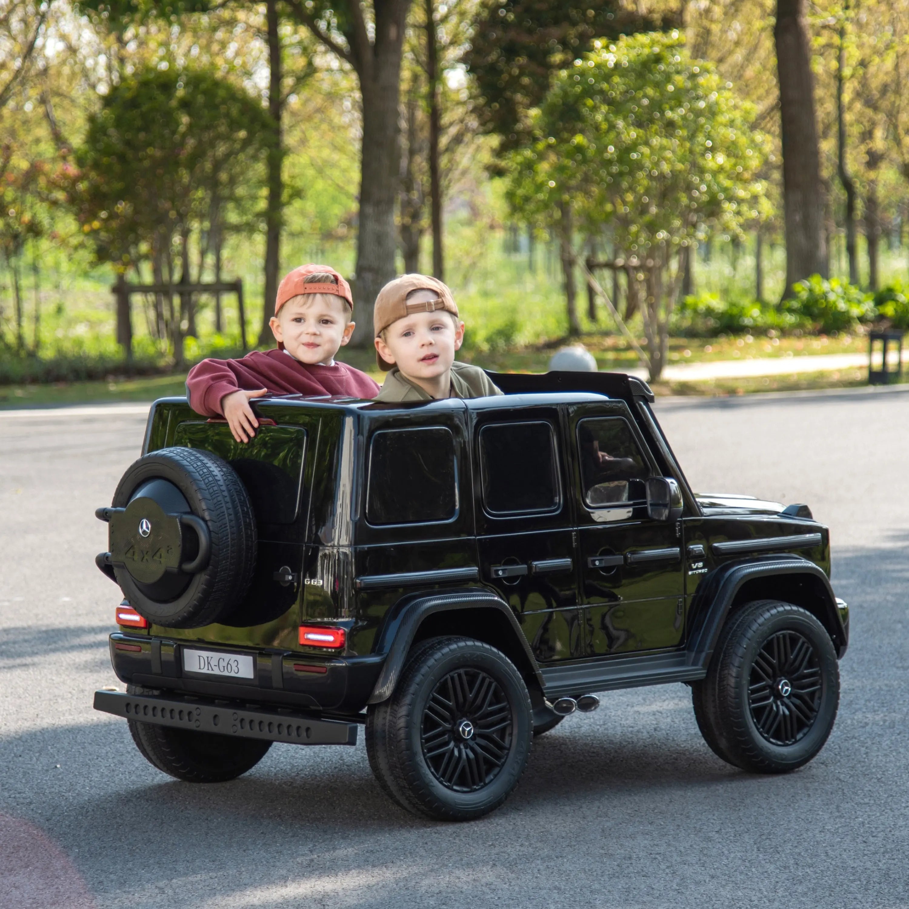 Two kids riding in a black 24V Mercedes-Benz G-Wagon toy car outdoors on a sunny day