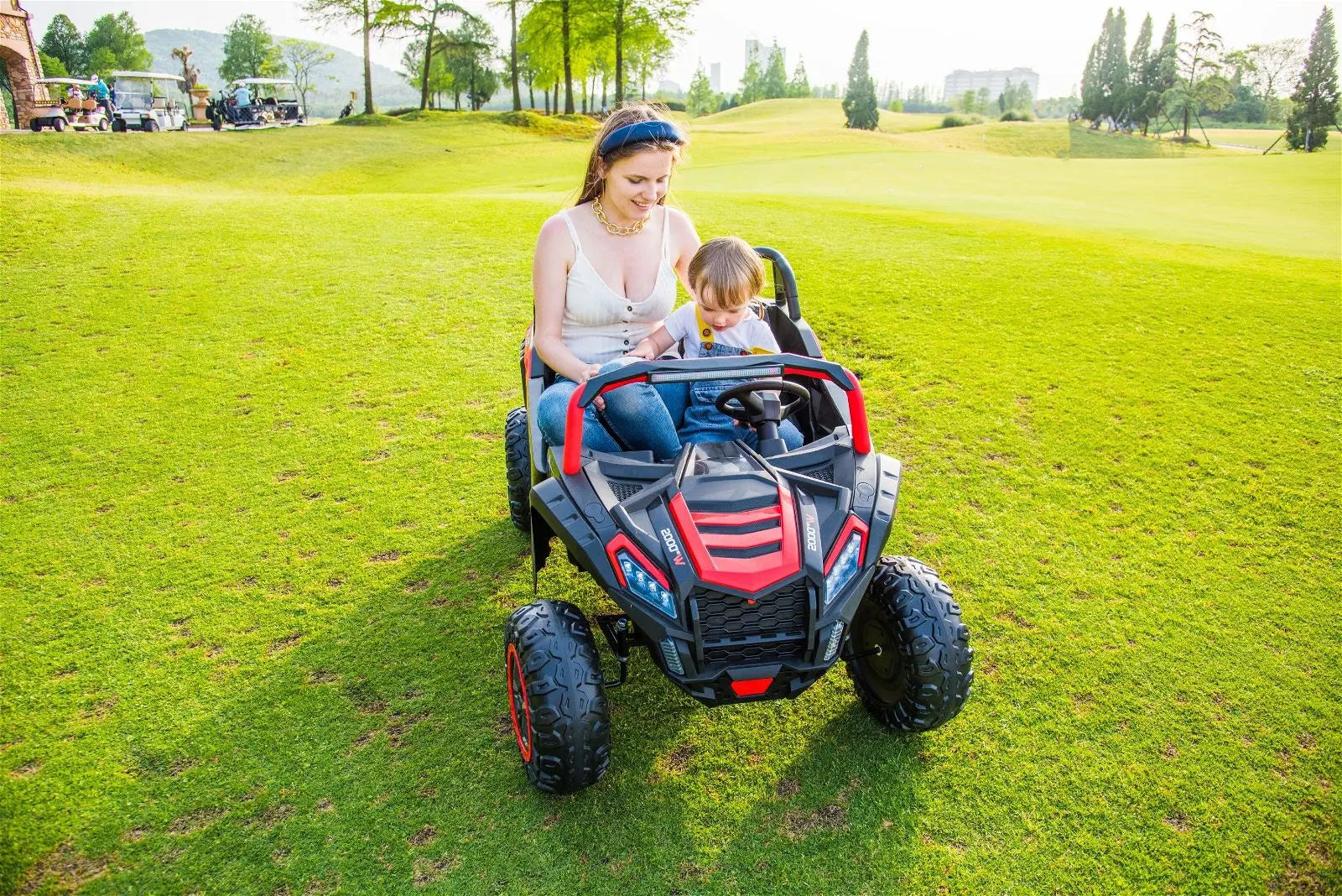 Woman and child riding a 2-seater 24V kids dune buggy on grassy outdoor field