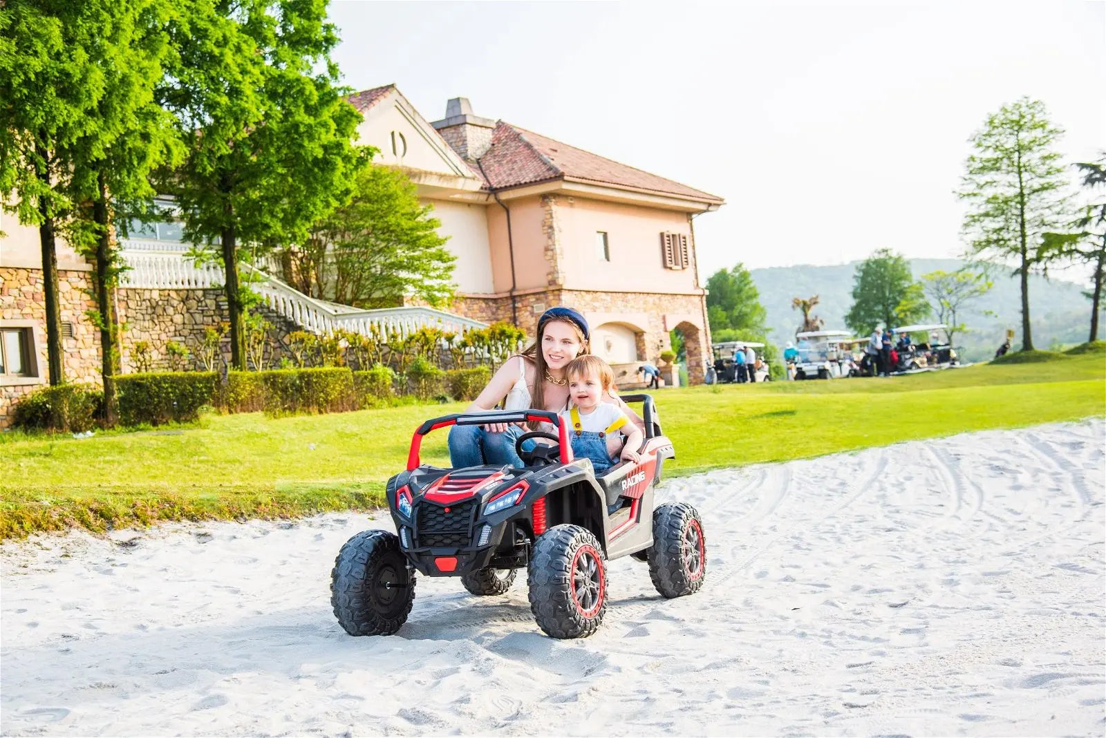 Two children riding a 24V 4x4 red dune buggy toy on sand in a sunny outdoor park.
