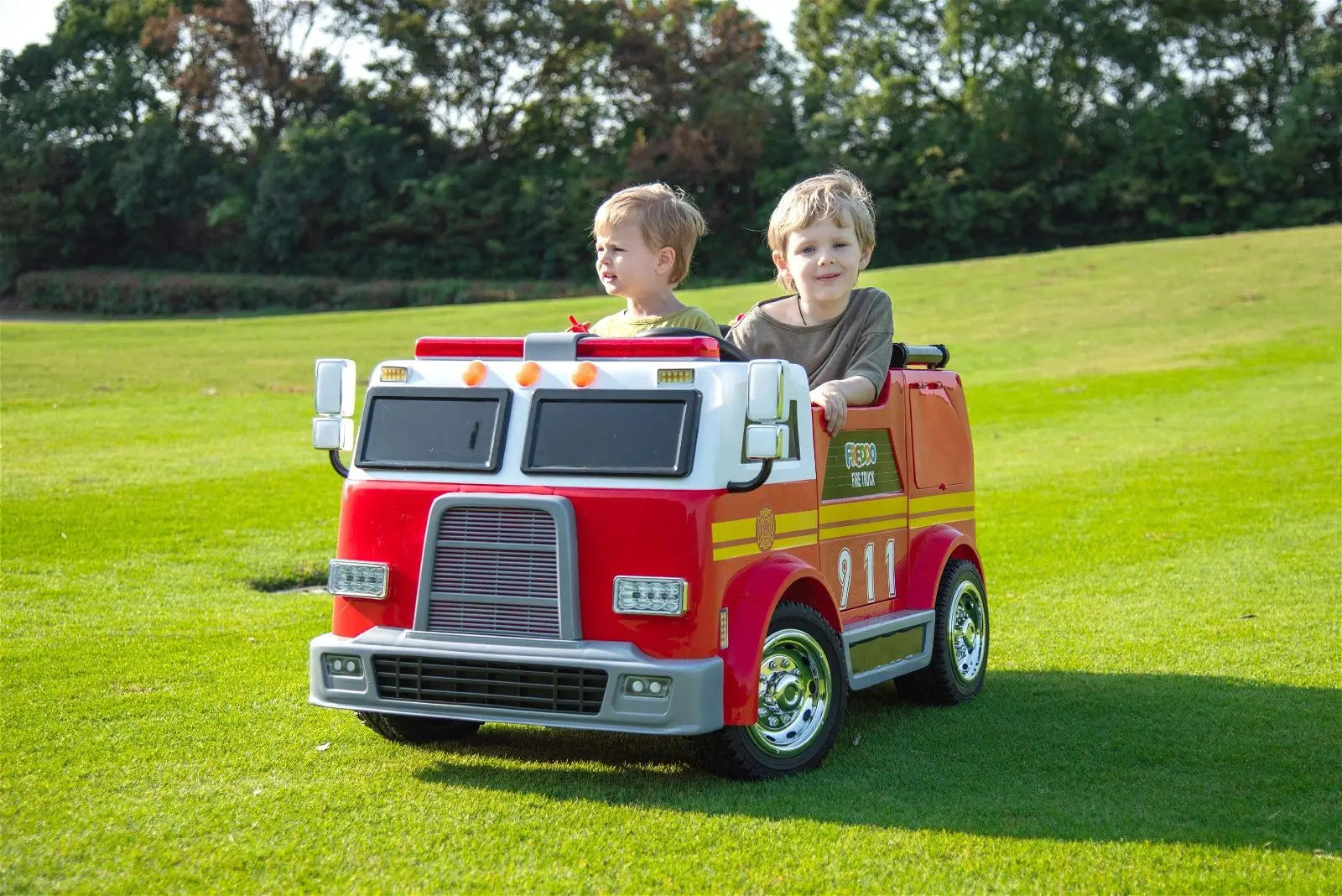 Two children riding a 2-seater red ride-on fire truck toy on green grass outdoors