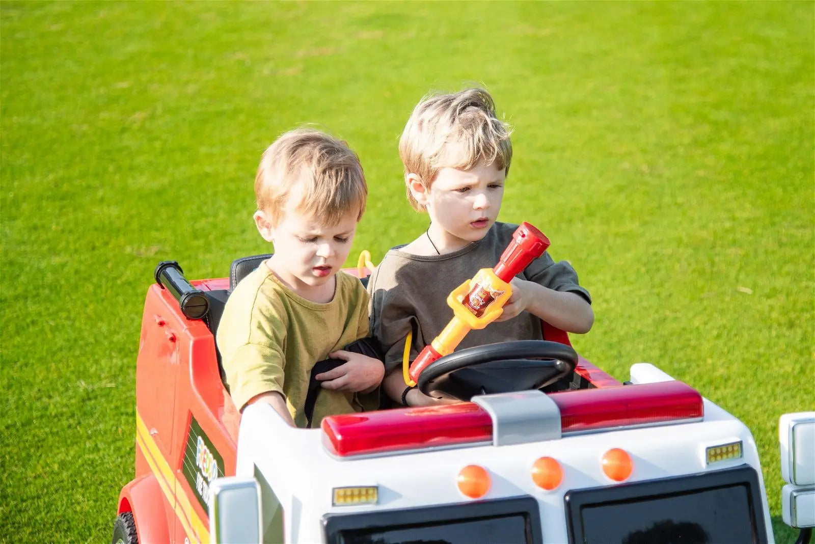Two kids riding a red 2-seater toy fire truck on green grass outdoors