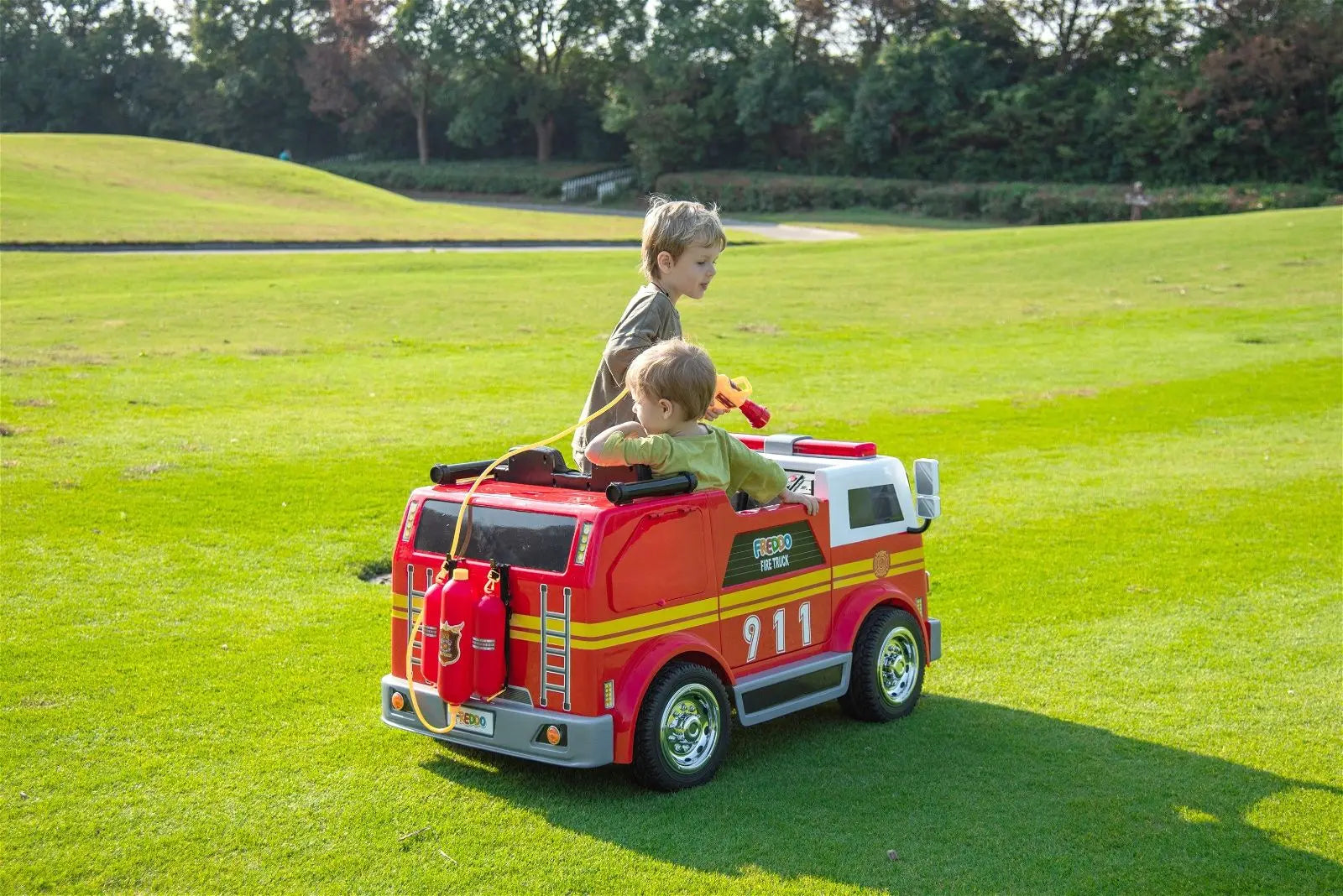 Two kids riding a red 24V electric fire truck toy on a grassy outdoor field