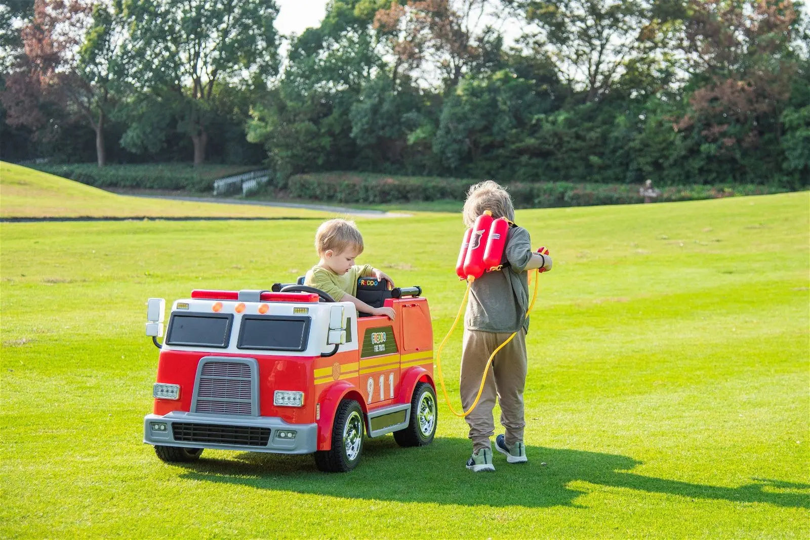 Two children playing on grass with a red 2-seater ride-on fire truck toy