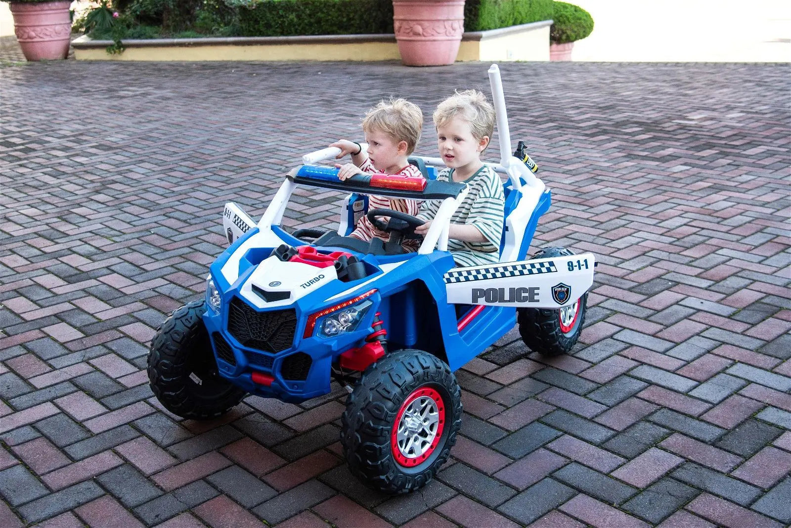 Two kids riding a blue and white police UTV toy car with lights and sirens on a paved driveway